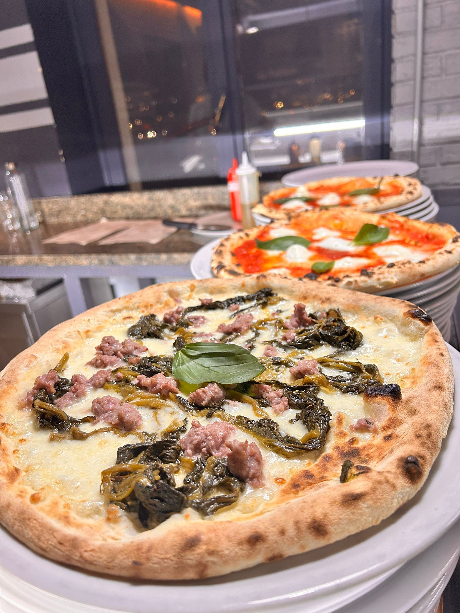 Tre pizze su piatti. La pizza in primo piano è condita con ingredienti diversi, le altre pizze sono con formaggio e basilico. Sullo sfondo, una finestra e un bancone.