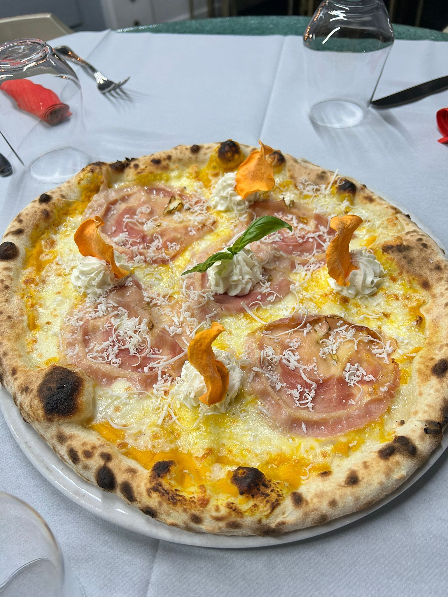 Pizza su un piatto bianco, con prosciutto, formaggio e guarnizioni arancioni, servita su una tovaglia bianca.