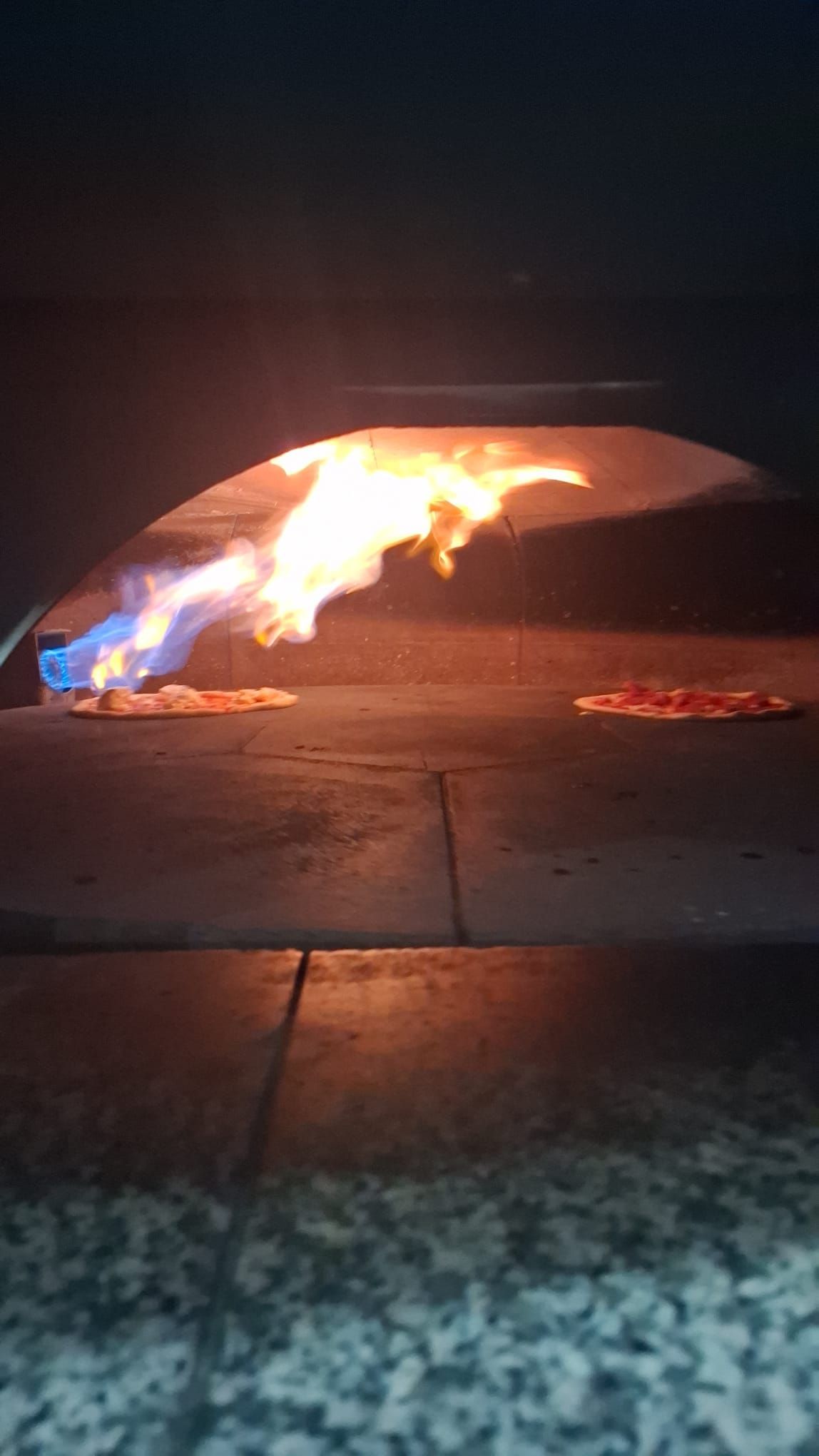 Cottura della pizza nel forno a legna con fiamme; le pizze sono visibili all'interno del forno in pietra.