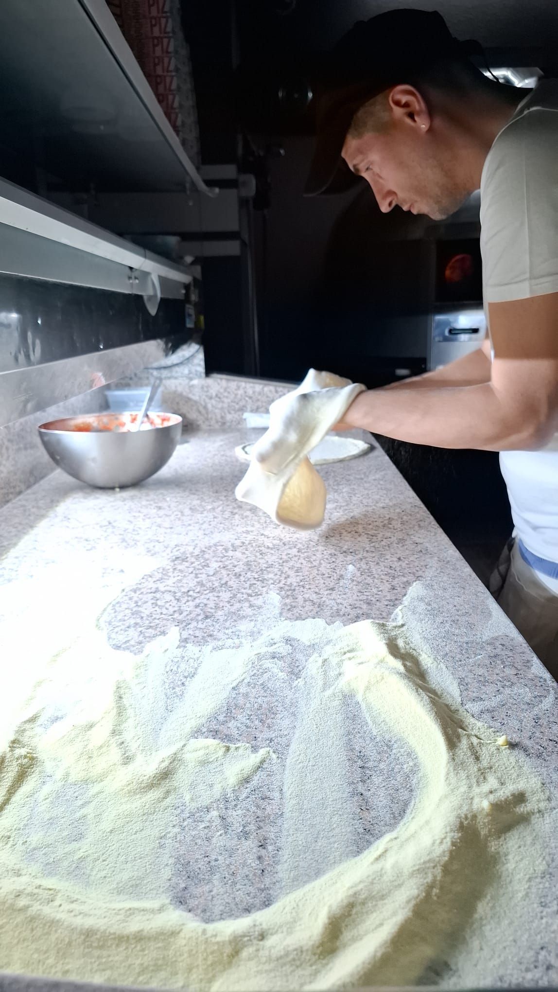 Persona che prepara l'impasto della pizza su un piano di lavoro infarinato, vicino a una ciotola di metallo con la salsa, in un ambiente interno.