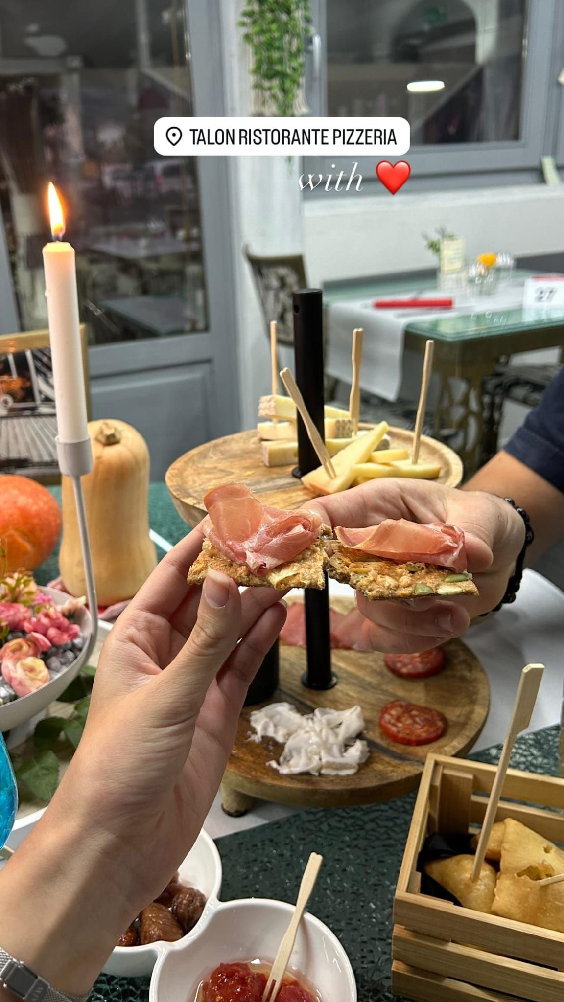 Mani che tengono cracker ricoperti di prosciutto, vicino a un banco di cibo a più livelli. Ambiente a lume di candela con antipasti.