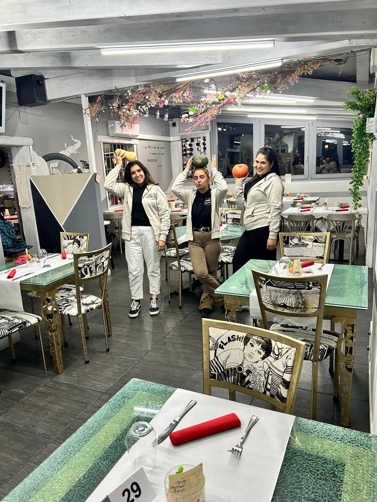Tre persone in posa in un ristorante con tavoli e sedie decorati, alzano le mani.