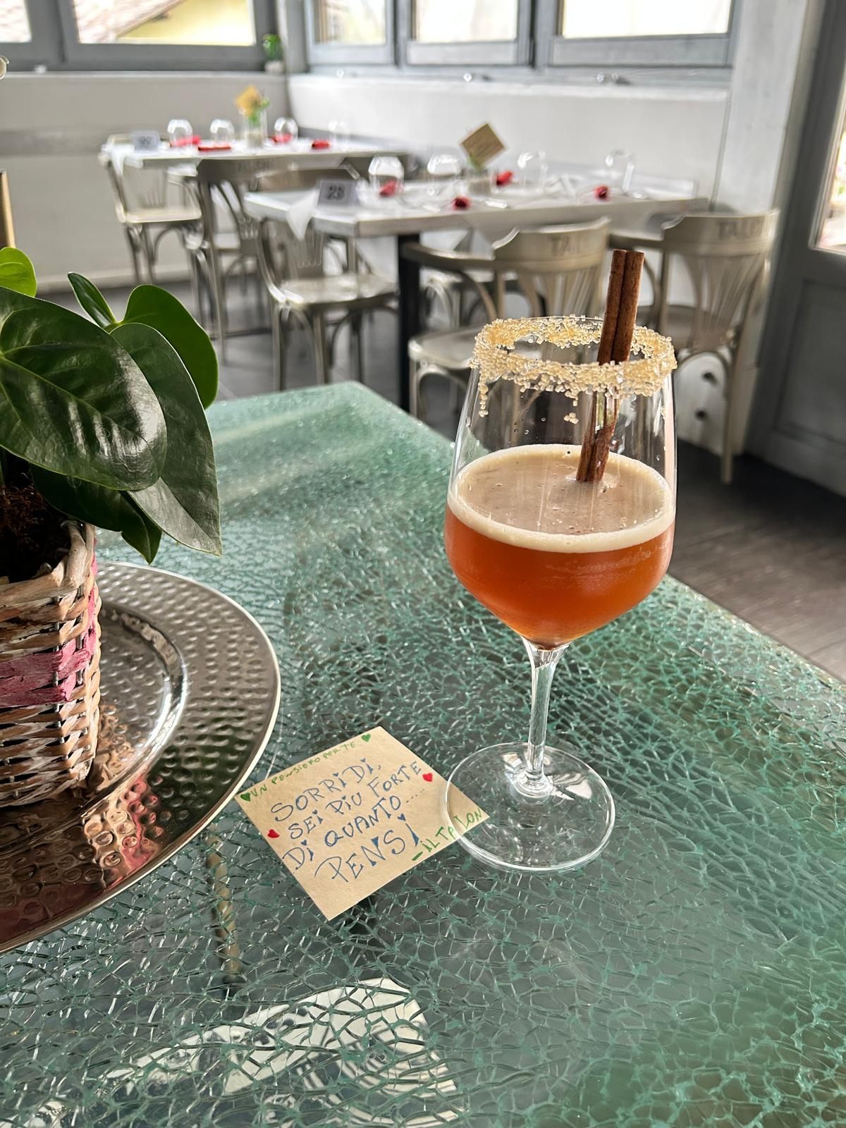 Cocktail con stecca di cannella, bordo di zucchero e un biglietto su un tavolo con ripiano di vetro in un ambiente da ristorante.