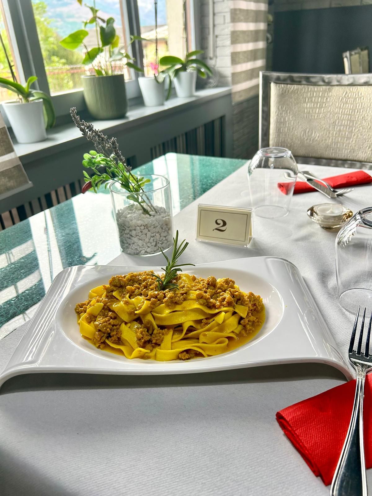 Piatto di pasta su un piatto bianco, tavola apparecchiata con tovagliolo rosso e finestra con piante sullo sfondo.