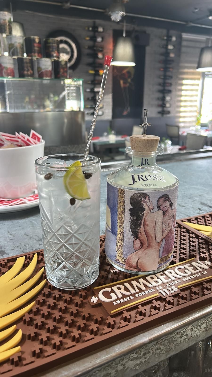 Bottiglia di cocktail e liquore su un bancone. Bicchiere alto con ghiaccio e lime. Bottiglia con un'opera d'arte raffigurante un nudo.