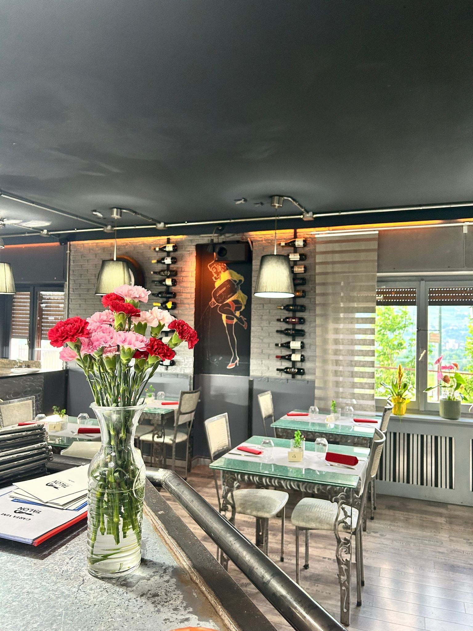 Interno di un ristorante con tavoli apparecchiati per cenare, fiori in un vaso e vista sugli alberi all'esterno.