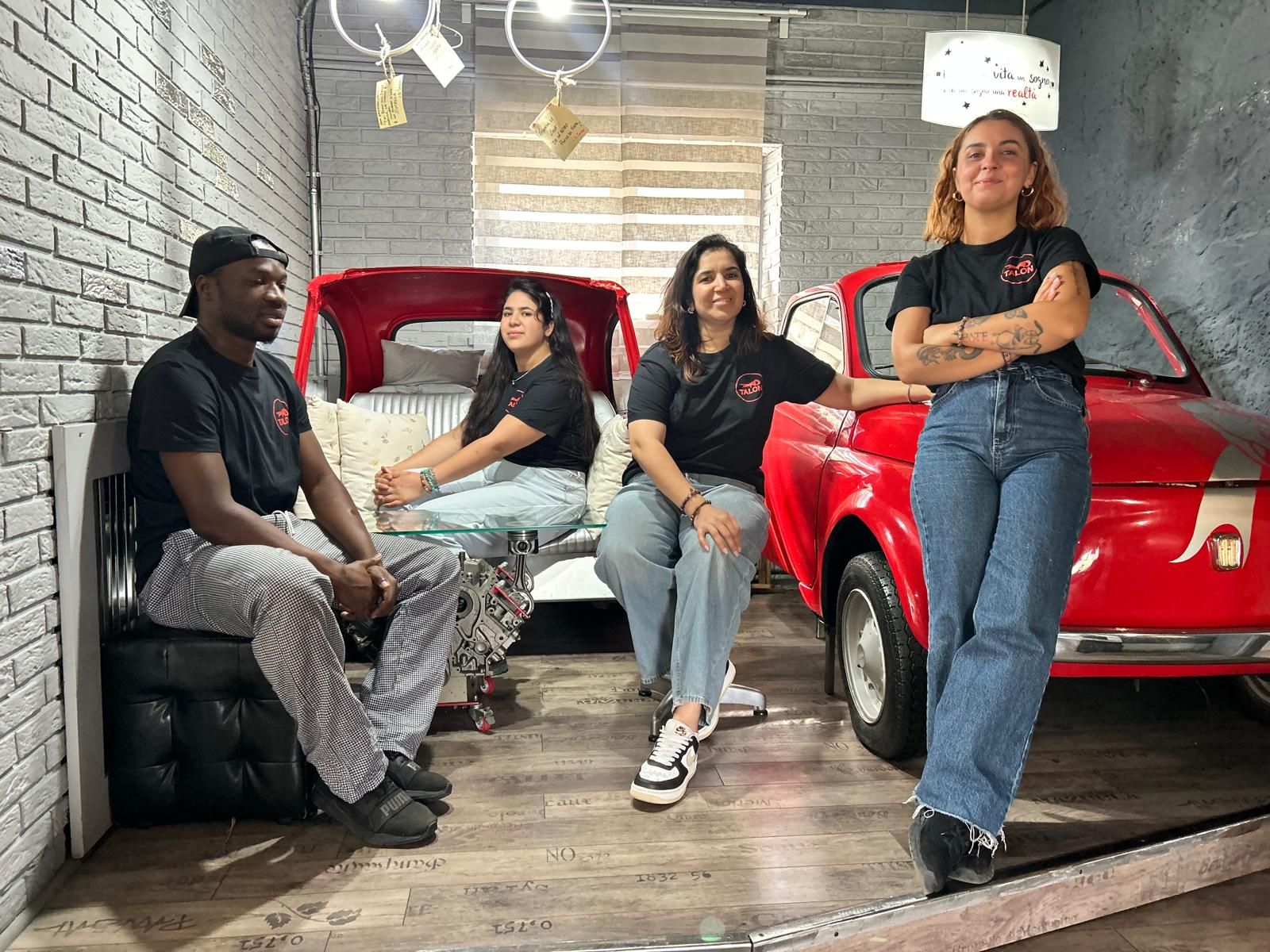 Quattro persone in una stanza con un'auto rossa; una persona è appoggiata all'auto, le altre sono sedute lì vicino.