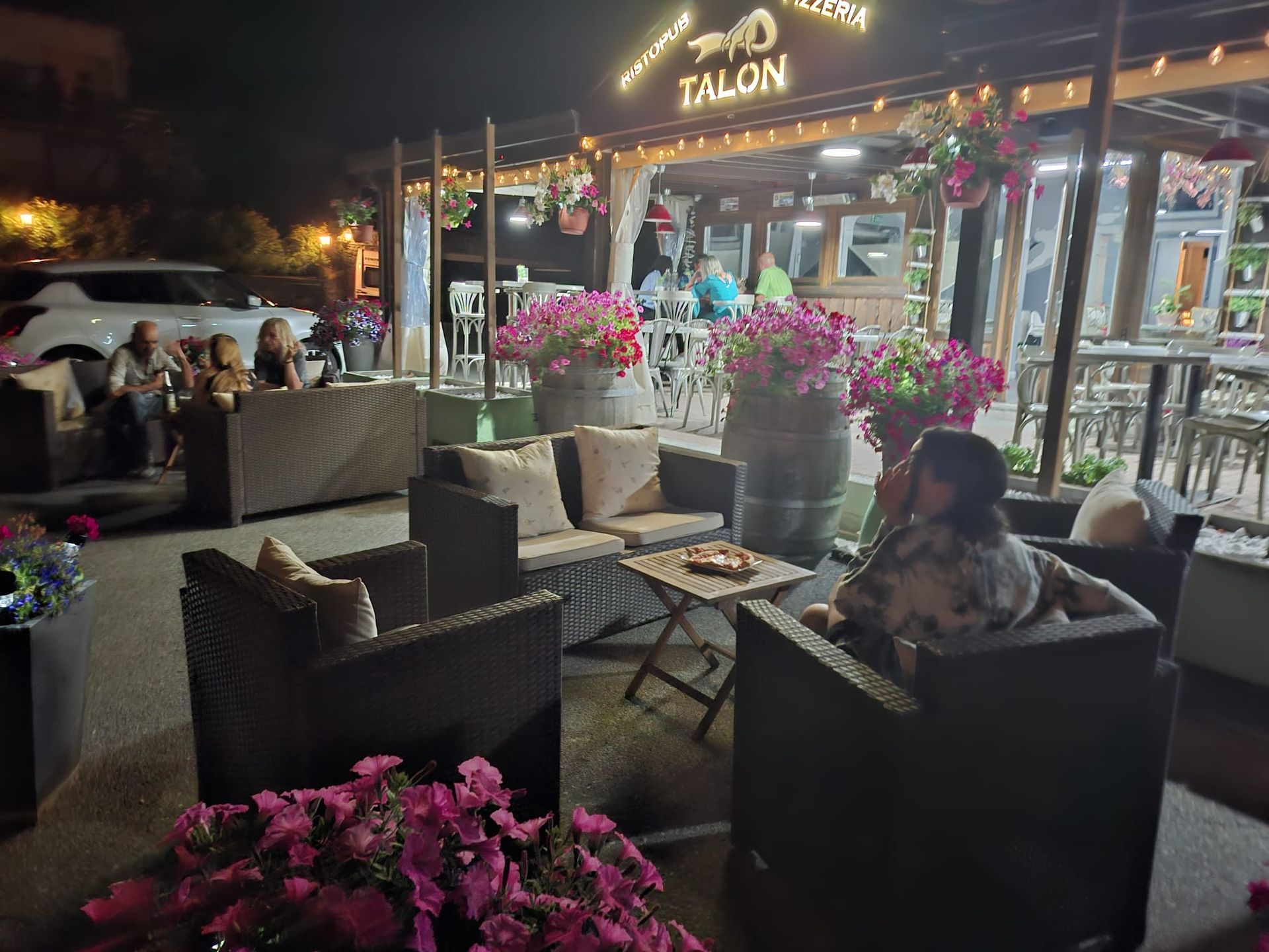 Patio esterno di sera, ristorante Talon. Persone sedute ai tavoli circondate da fiori e luci a ghirlanda.