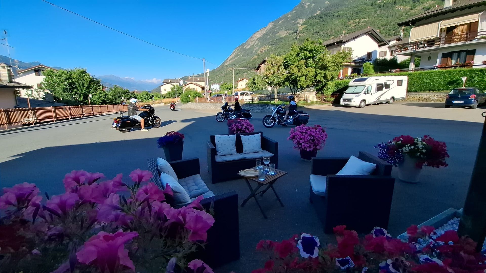 Area salotto all'aperto con fiori rosa, motociclette parcheggiate e montagne sullo sfondo.