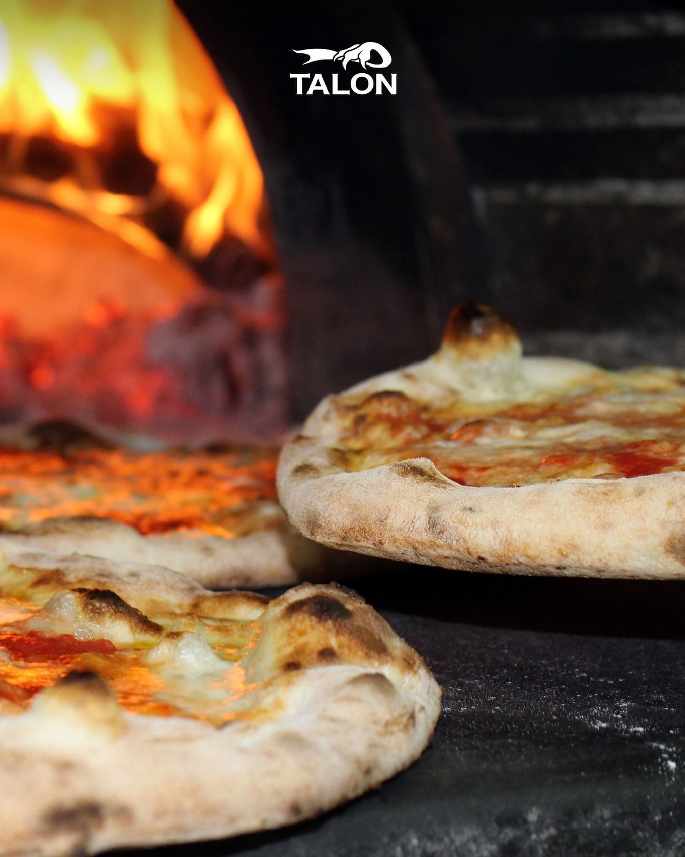 Pizze cotte in un forno a legna con fiamme visibili sullo sfondo.