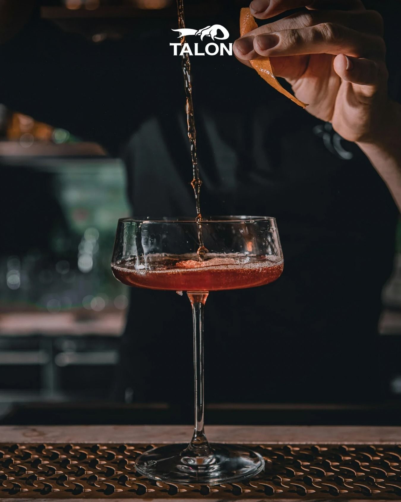 Cocktail versato in un bicchiere con stelo, ambientazione da bar. Liquido scuro, mani del barista visibili.