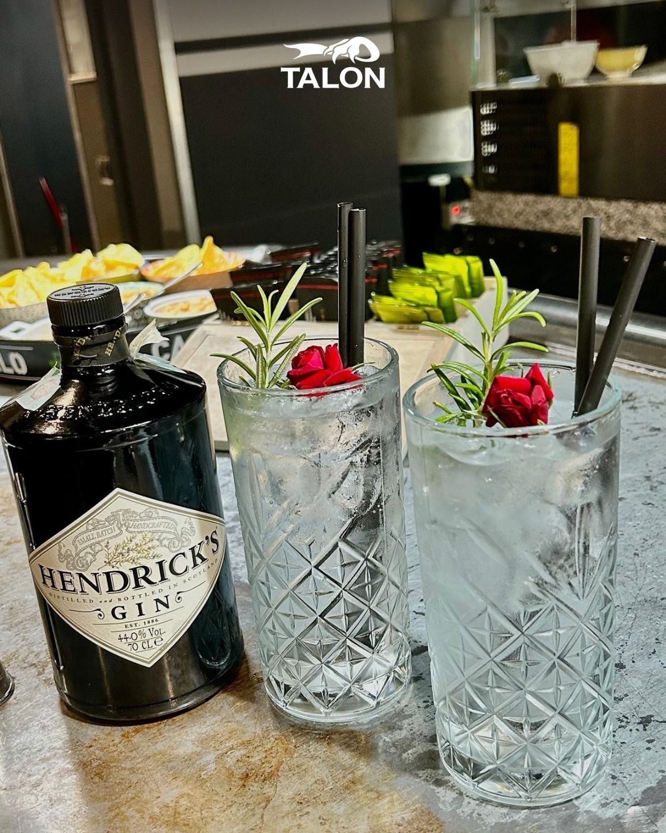 Bottiglia di gin Hendrick's con due cocktail, guarnita con rosmarino e fiori rossi, su un bancone del bar.