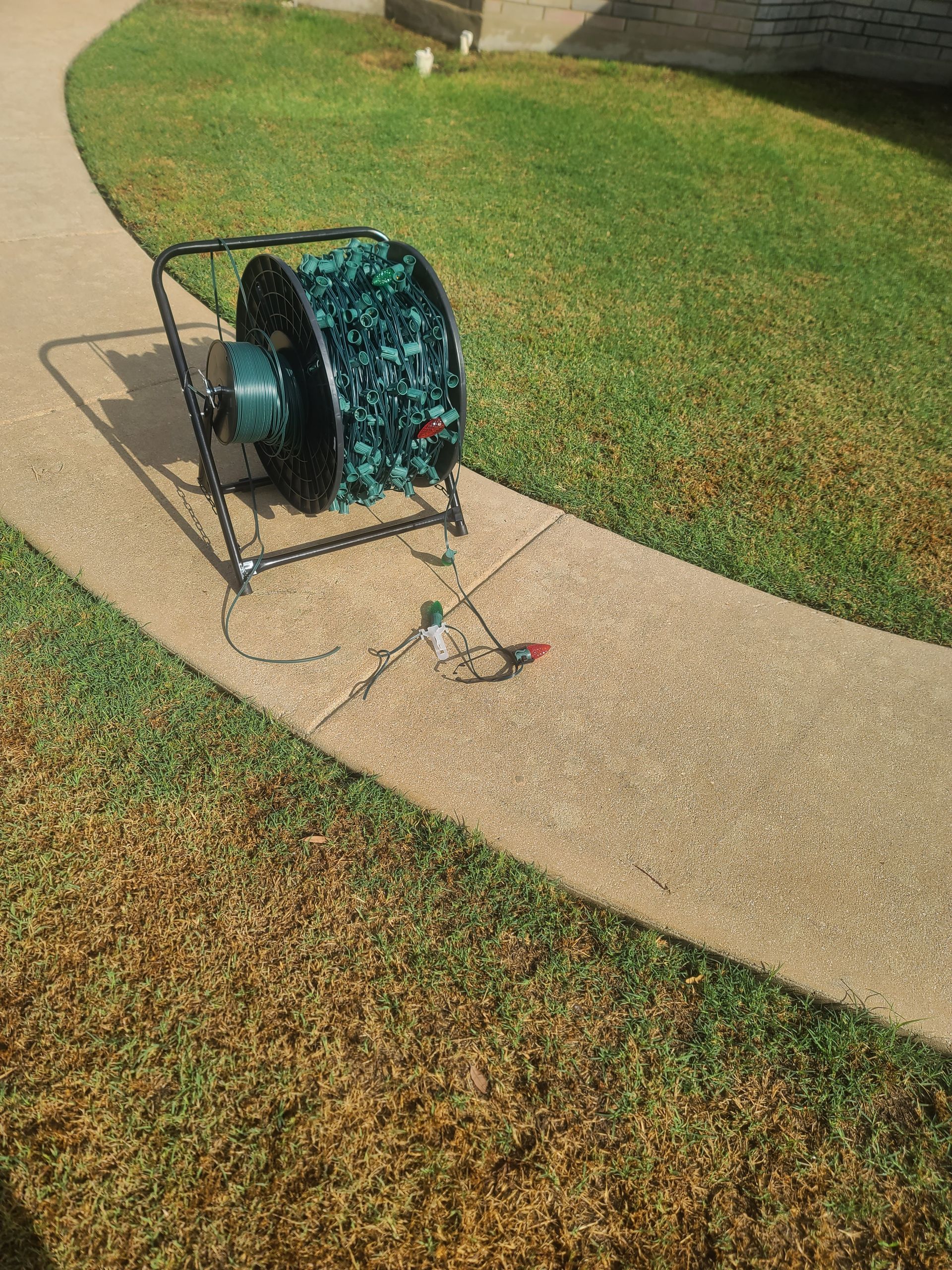 A large spool of green Christmas lights on a black stand, on a sidewalk next to grass.