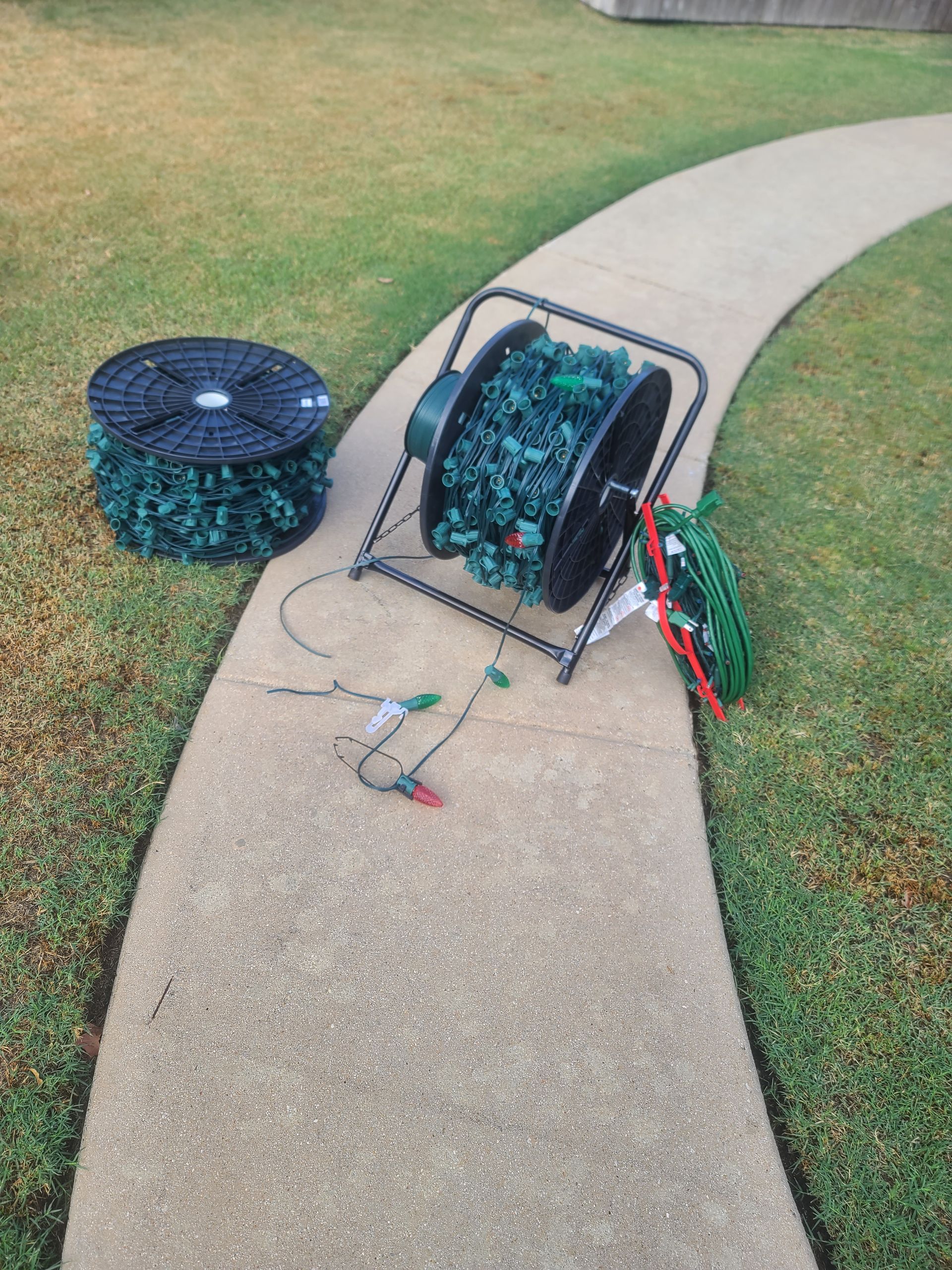 Spools of Christmas lights, one on a metal stand, on a curved sidewalk surrounded by grass.