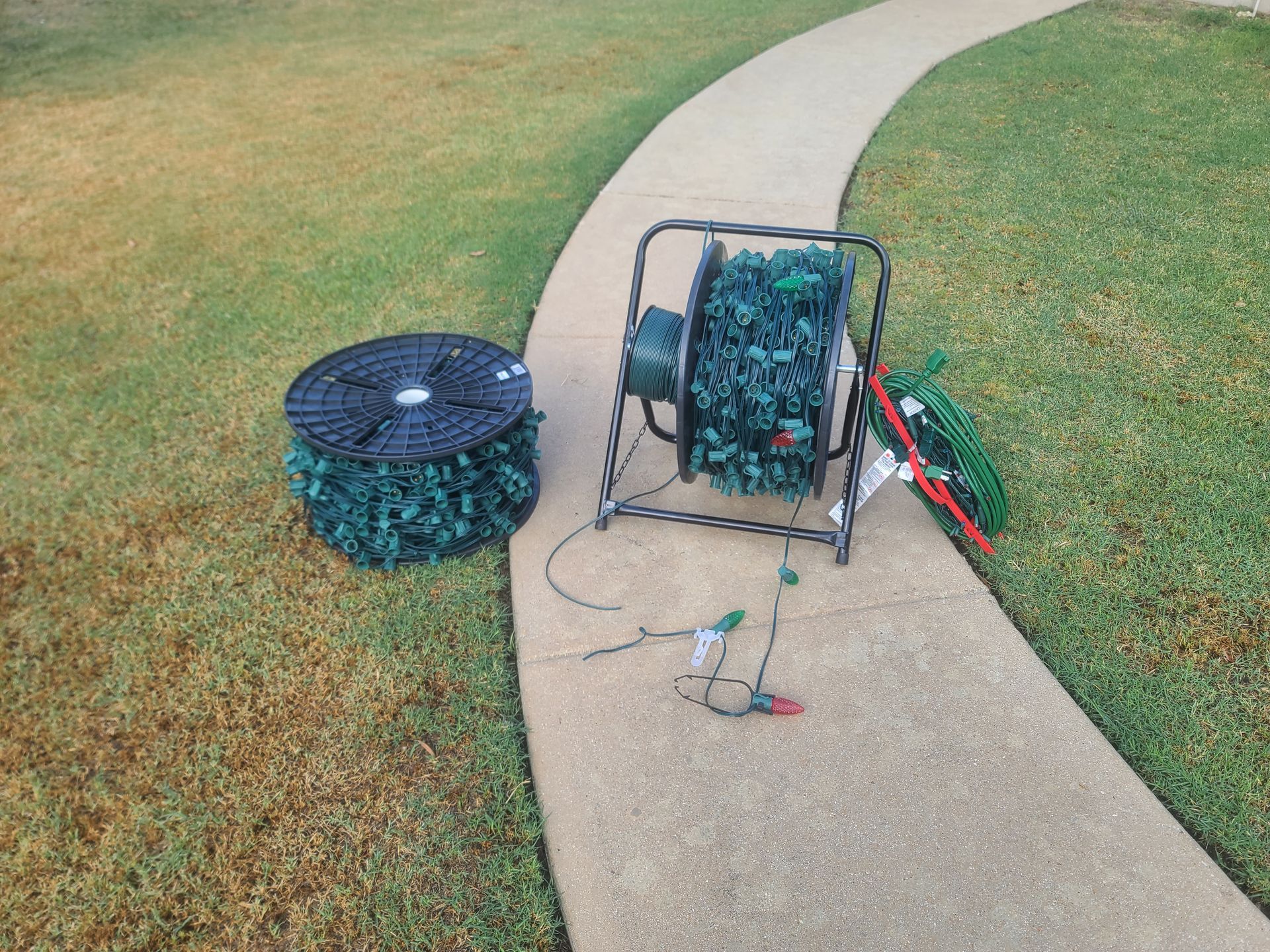 Spools of green garland on a sidewalk next to grass. One spool has a metal stand, and the other lies flat.
