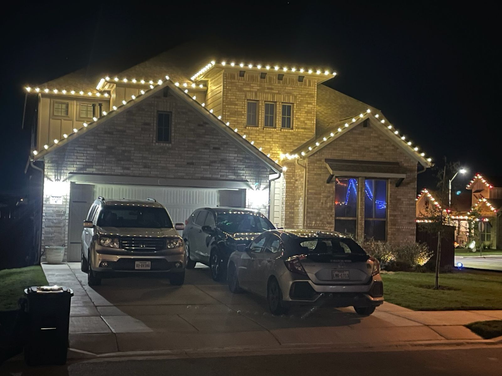 House decorated with white Christmas lights; cars parked in the driveway at night.