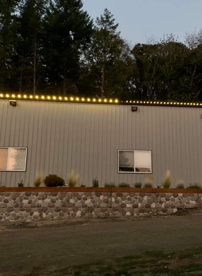 Building exterior with lit string lights along the roofline; stone and plant border below.