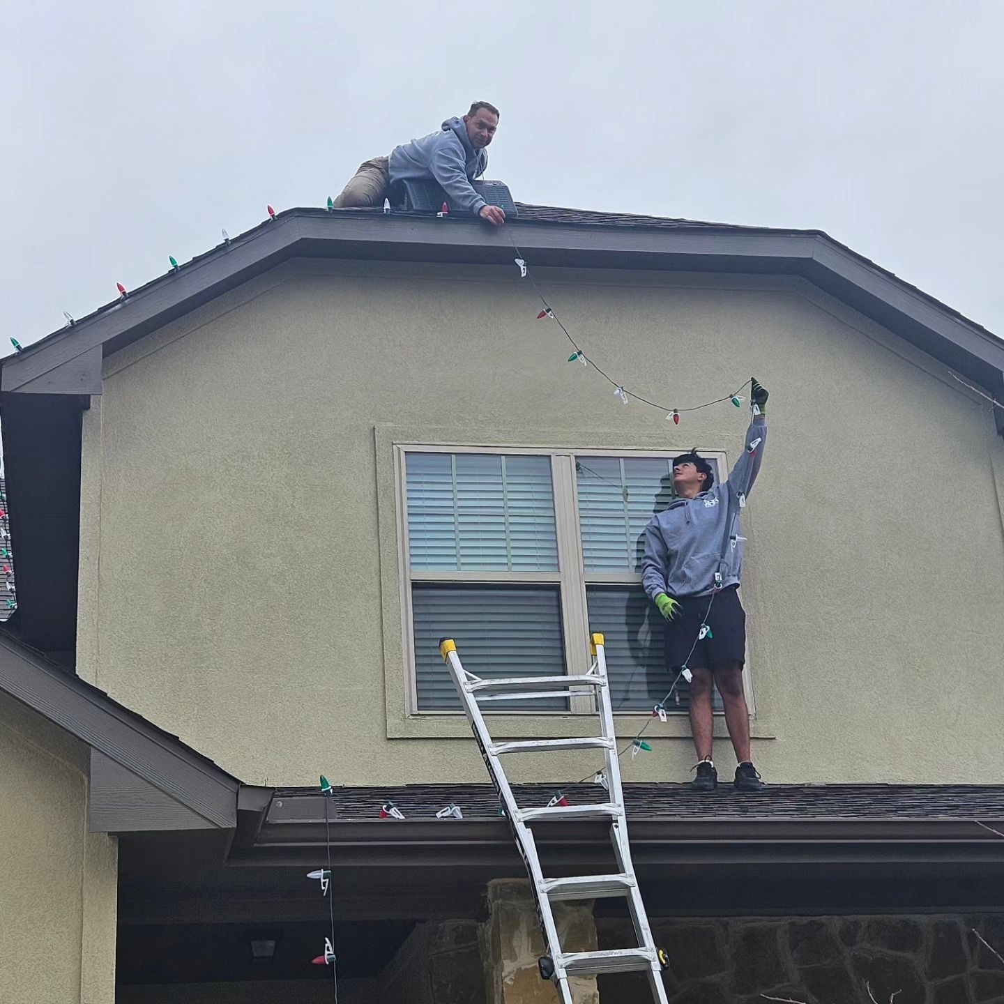 Two men install Christmas lights on a house roof. One on the roof, the other on a ladder.