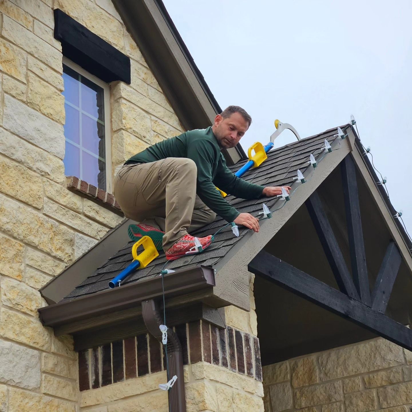 Man on roof attaching holiday lights with a yellow tool. House with stone and brown roof. Cloudy sky.
