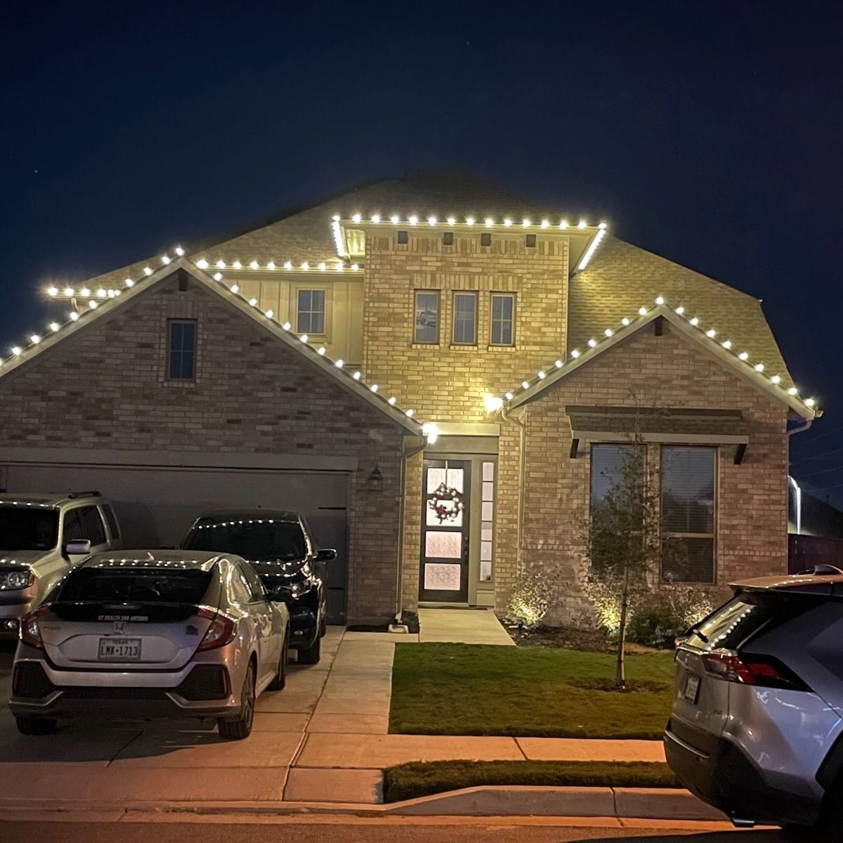 House decorated with white Christmas lights at night. Cars in driveway.