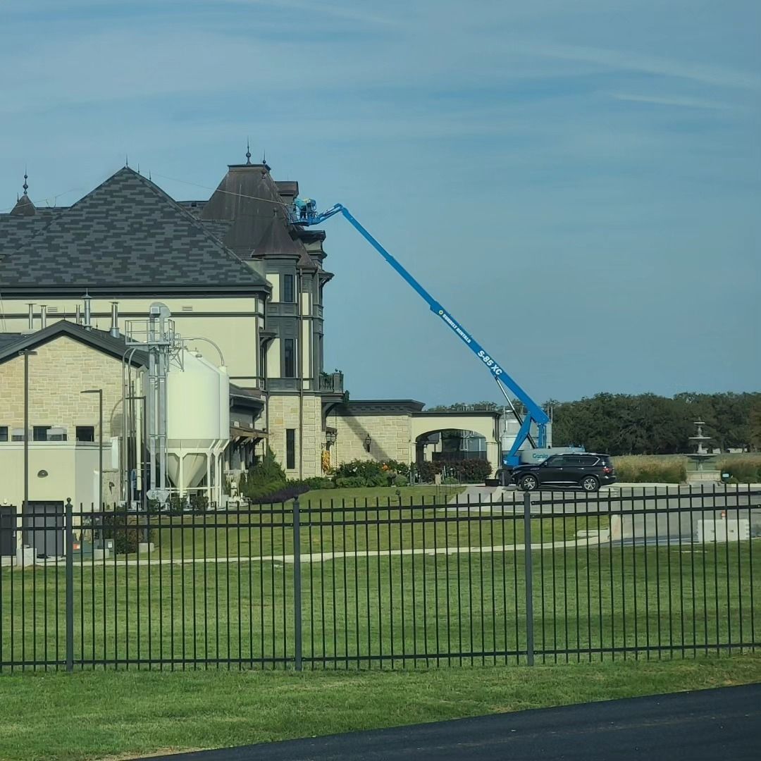 Blue boom lift reaching toward a beige building with a dark roof. A black SUV is parked nearby.