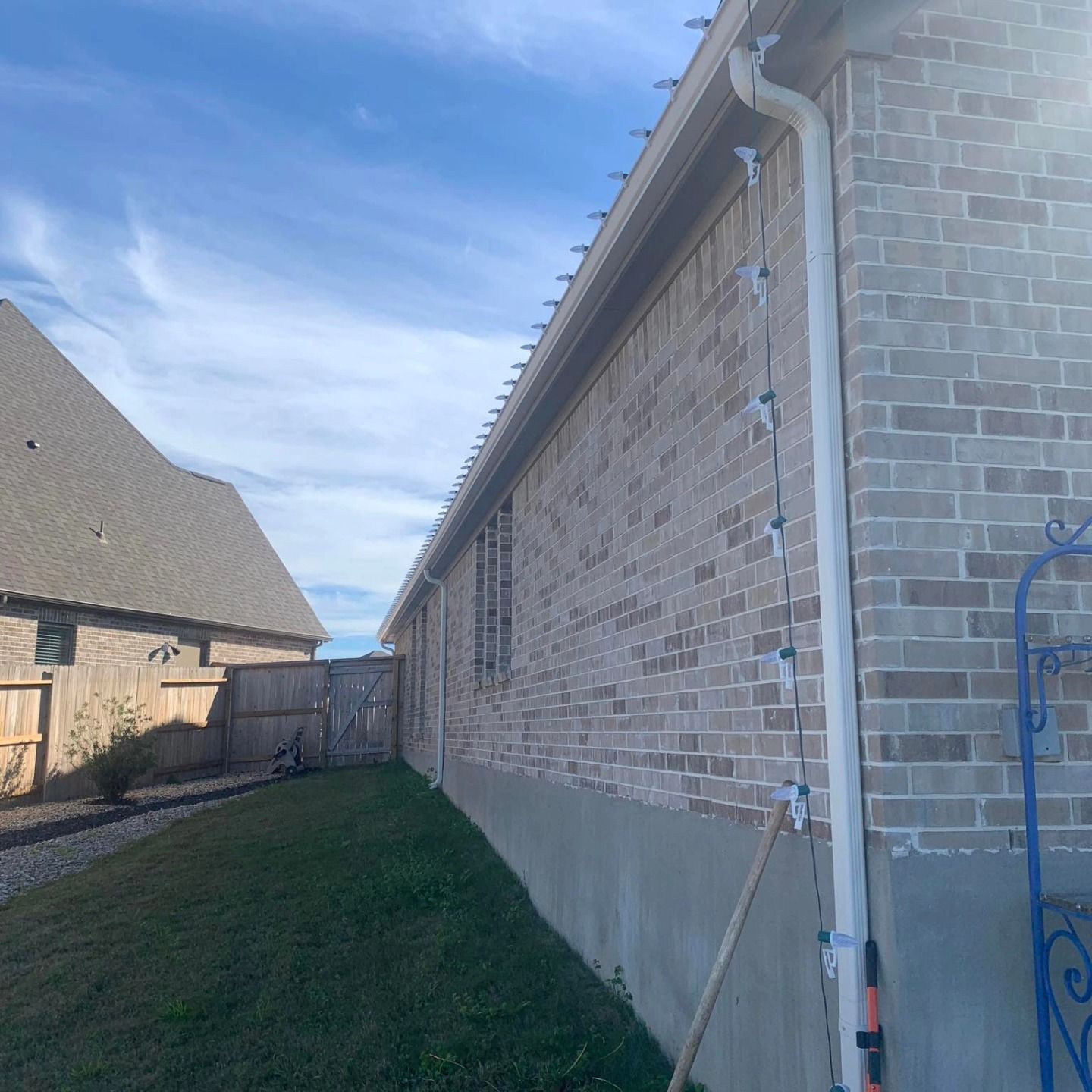 Brick house with gutter and lights, next to a yard with a fence. Blue sky in background.