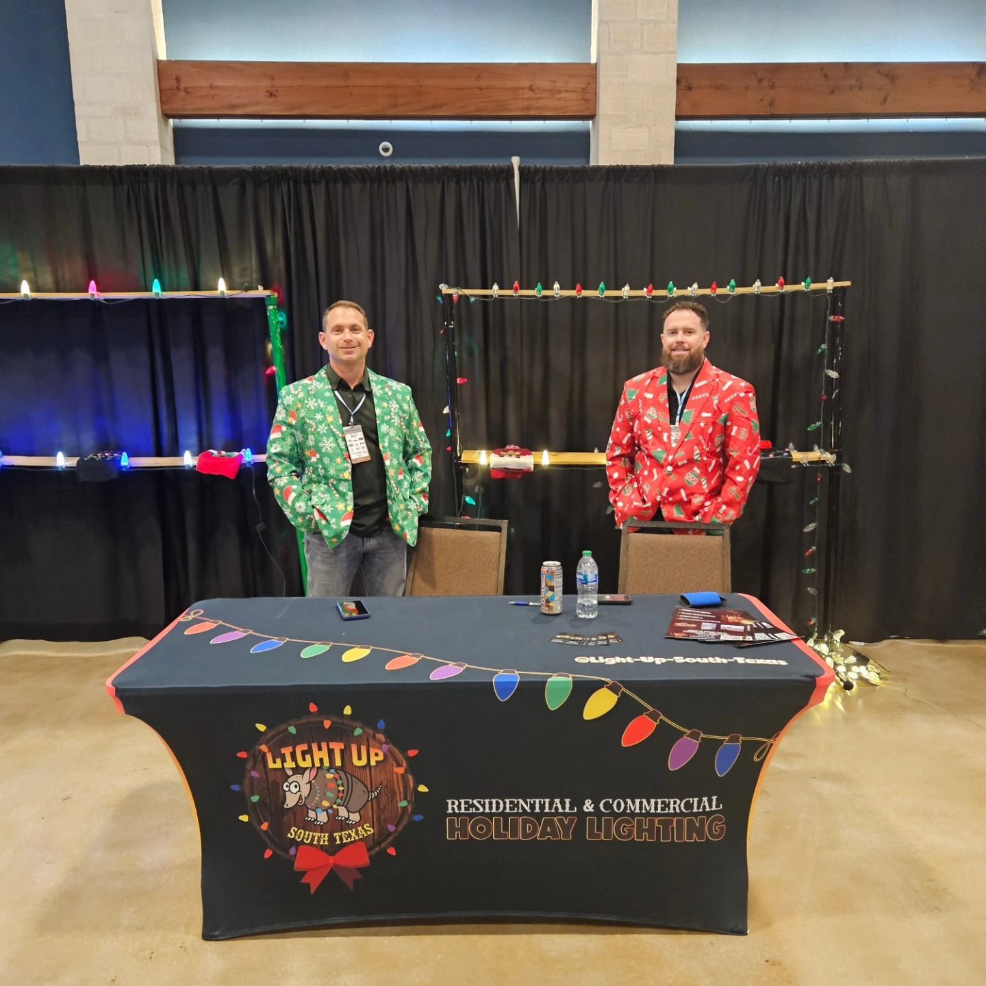 Two men in holiday sweaters stand at a booth advertising 