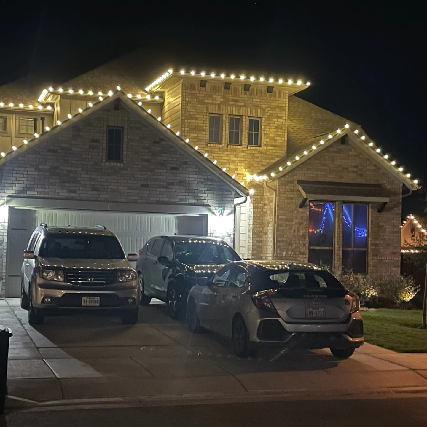 A house at night with holiday lights, and cars in the driveway.