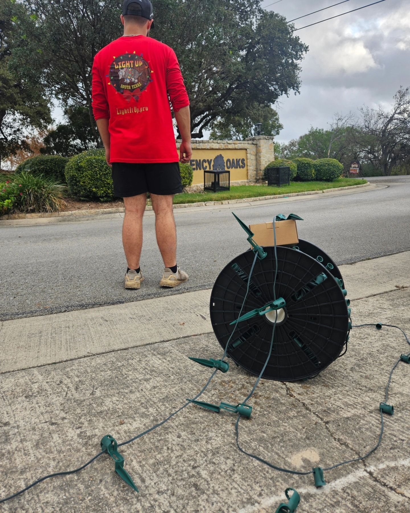 Man in red shirt and black shorts next to a large spool of green string lights on pavement.