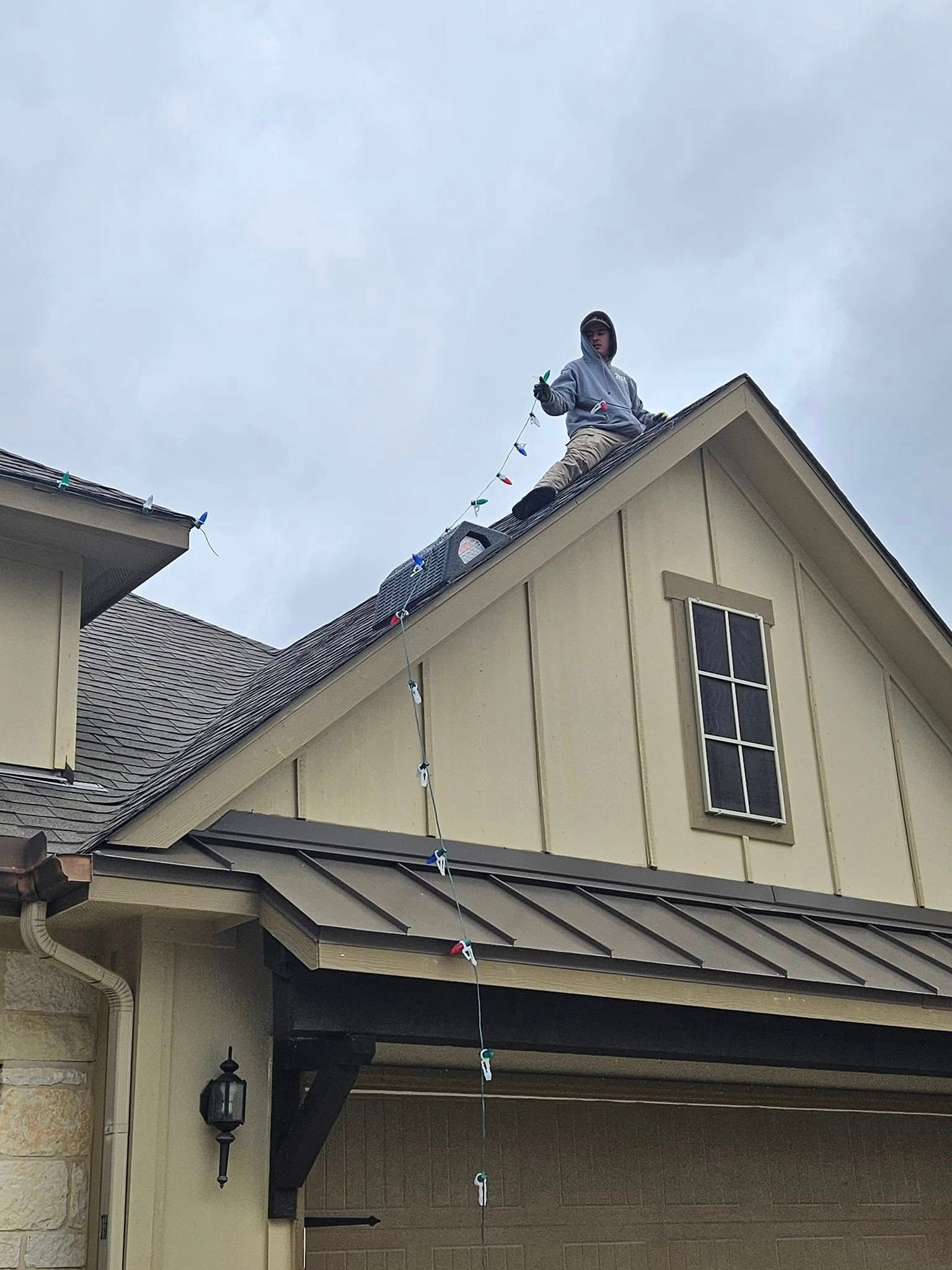 Person on roof installing lights on a house with dark gray roof and light tan siding.
