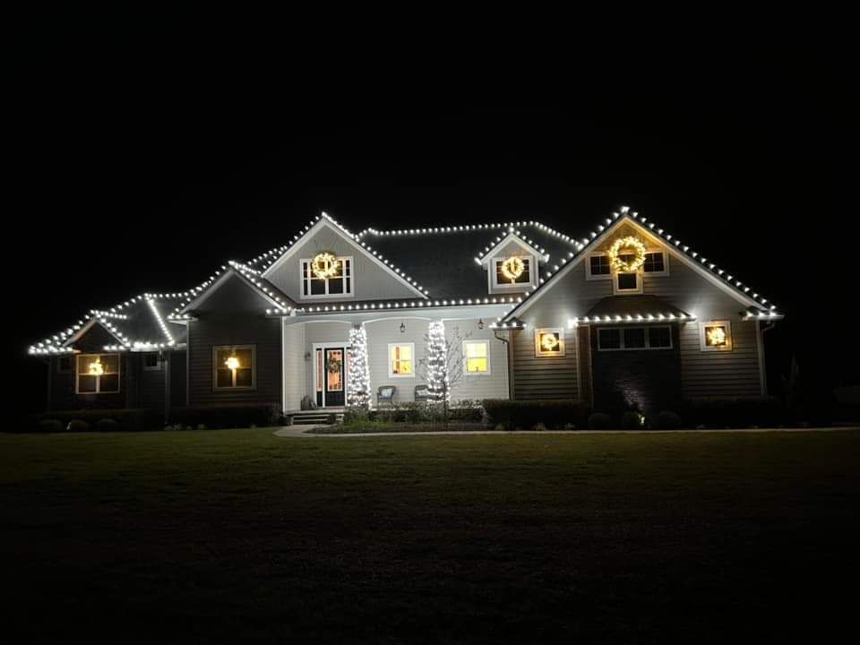 House at night, lit with white Christmas lights on roof, wreaths, and windows.