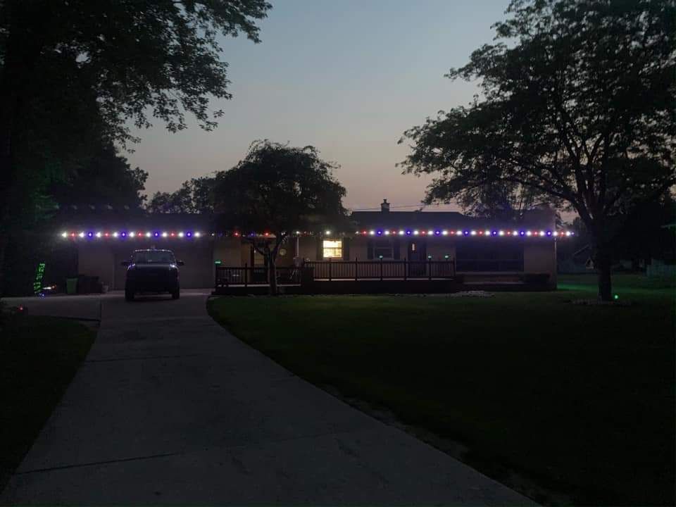 House at dusk with blue and white string lights along the roof; car in driveway.