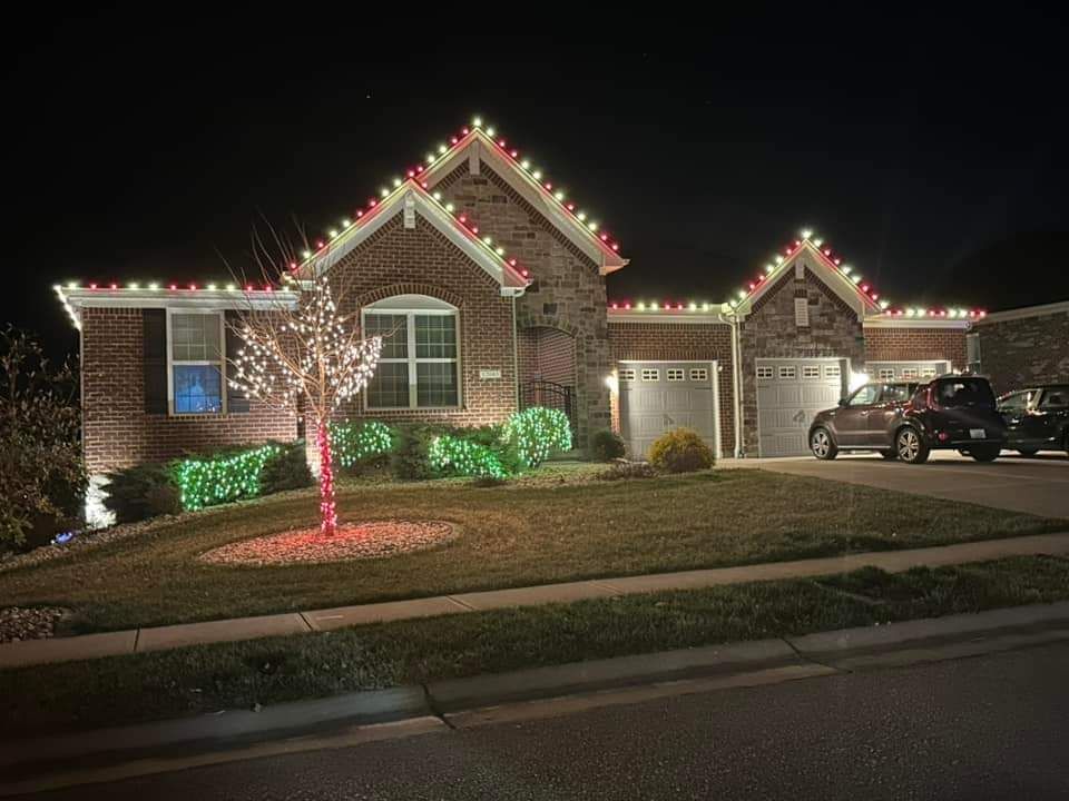 A brick house at night, decorated with Christmas lights on the roof and landscaping. A car parked in the driveway.