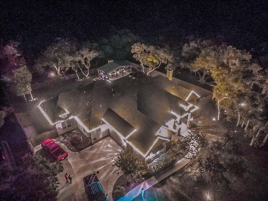 Night aerial view of a house with Christmas lights, red car in driveway, surrounded by trees.