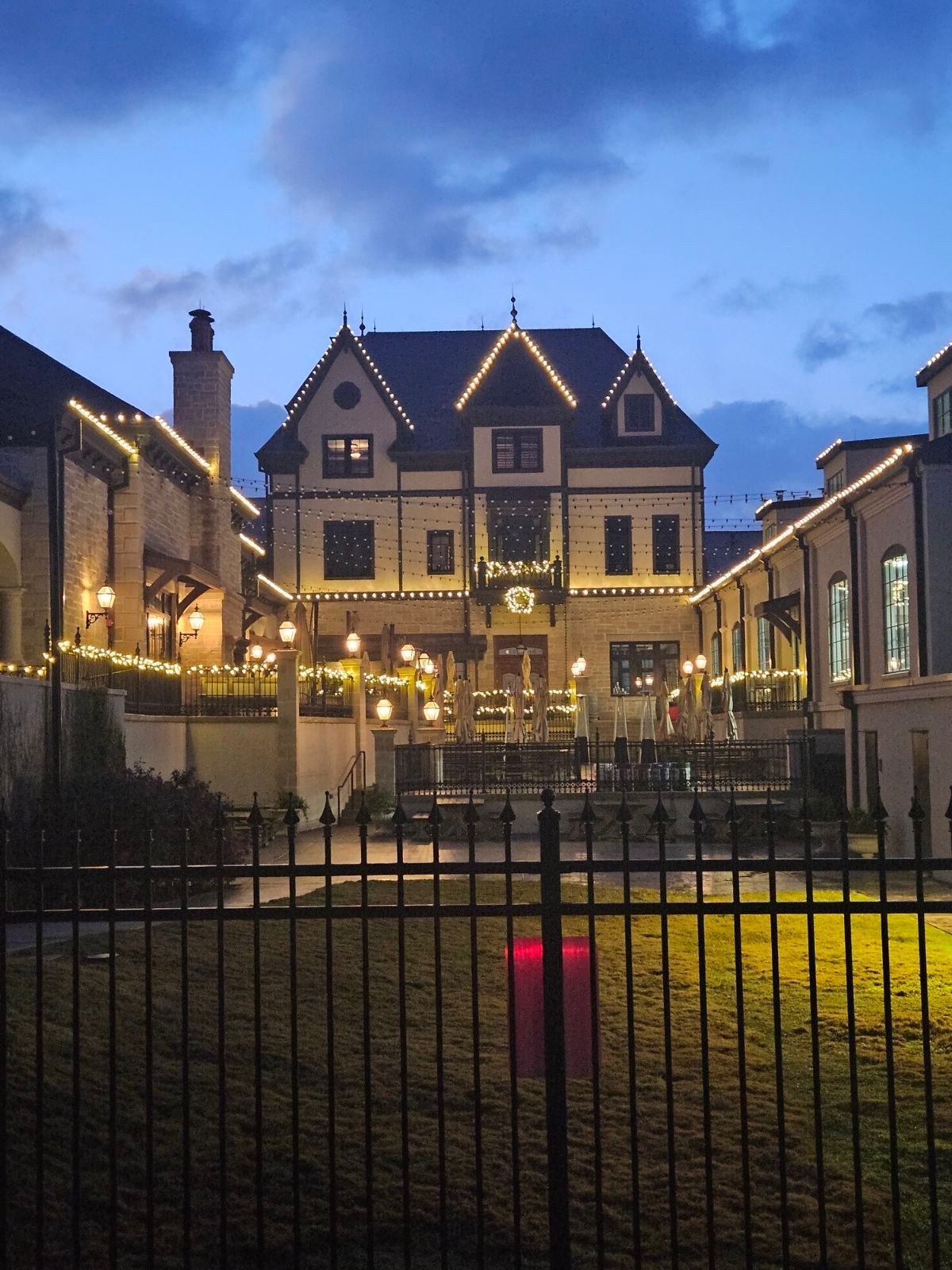 Night view of a building with lights, behind a black fence, under a cloudy sky.
