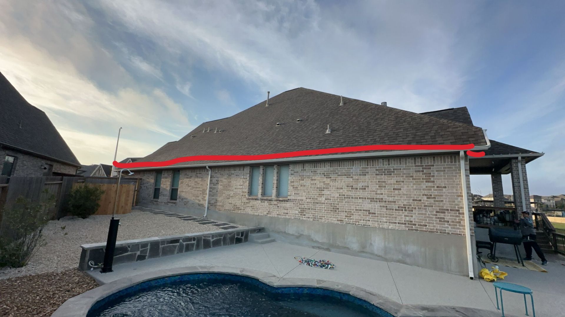 Brick house with dark roof, a pool, and people on the right side near a patio.