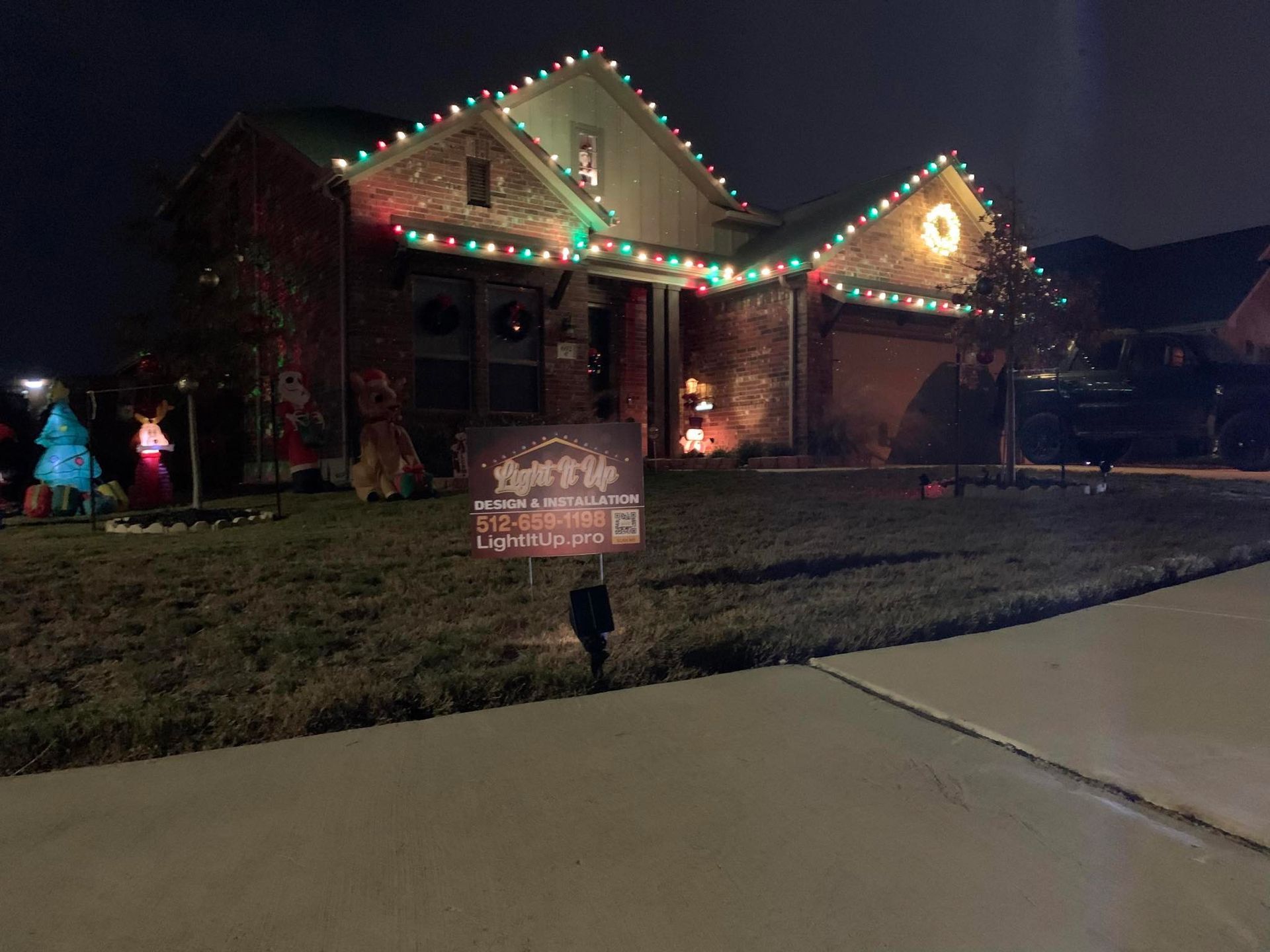 House decorated with red, green, and yellow Christmas lights. Lawn sign in front. Night setting.