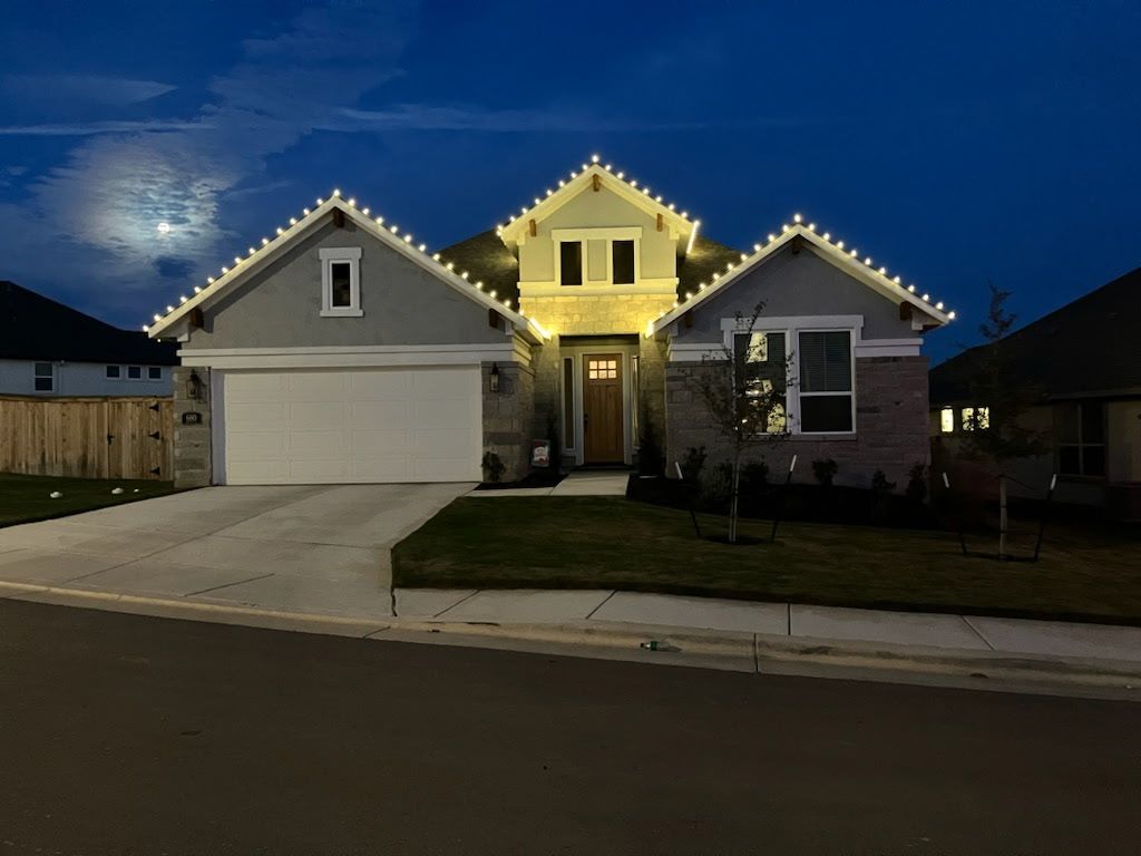 House at night, white lights outlining roof. Gray stucco and brick exterior, lit doorway, green lawn.