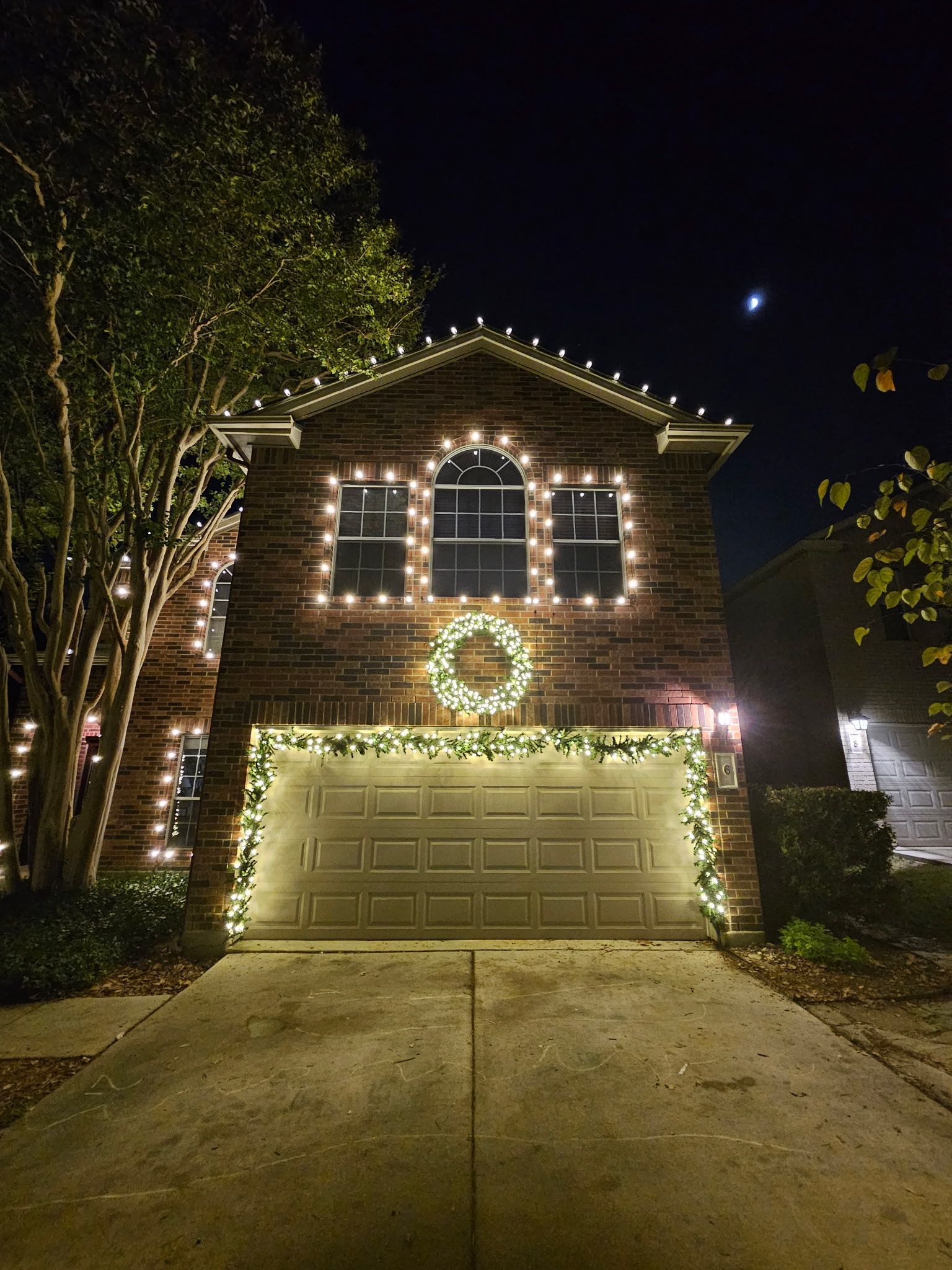 Two-story brick house lit with Christmas lights; garland around garage, windows, and roofline; wreath over the door.