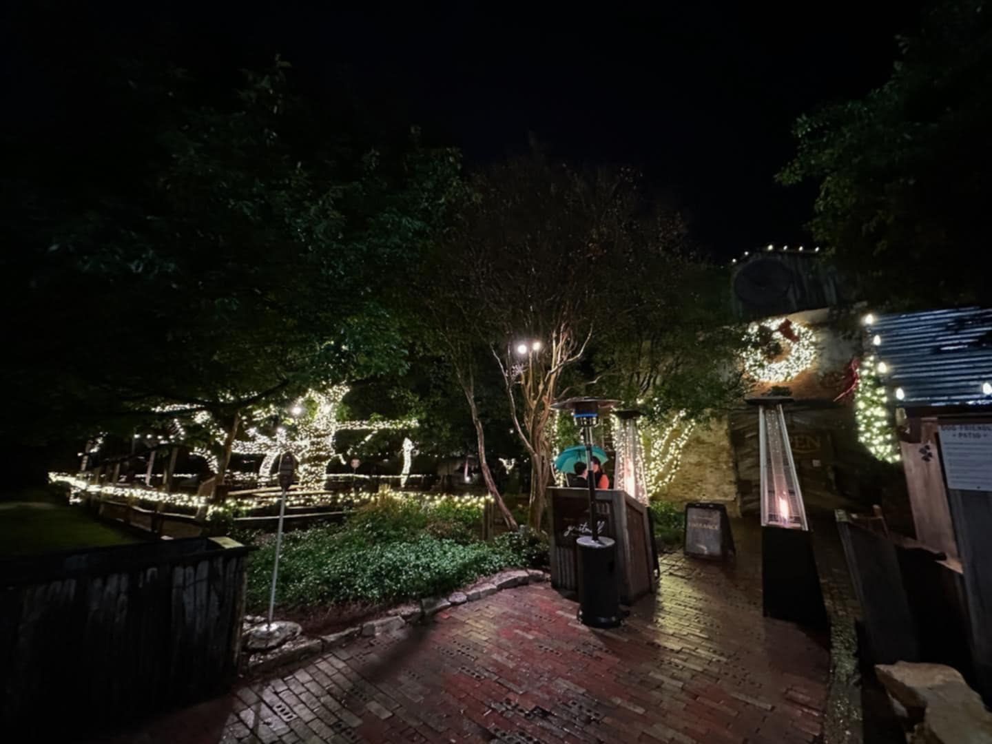 A night scene of a lit-up outdoor restaurant with trees, brick walkway, and twinkling lights.