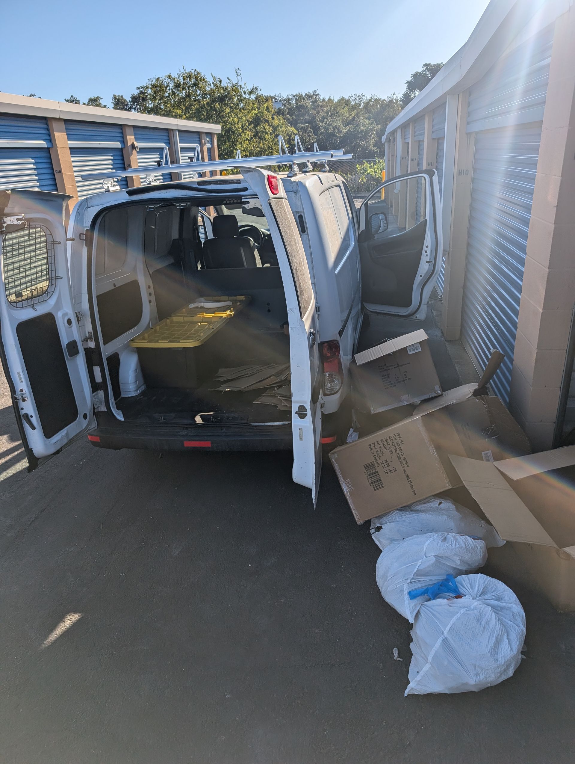 White van with open doors parked between storage units, unloading boxes and bags.
