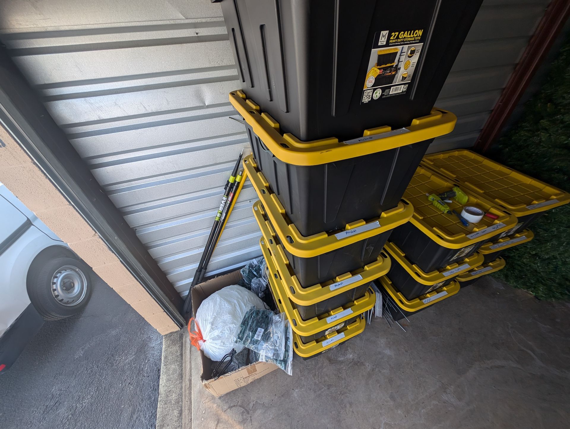 Storage unit interior with stacked black and yellow bins, cardboard box, and a white van.