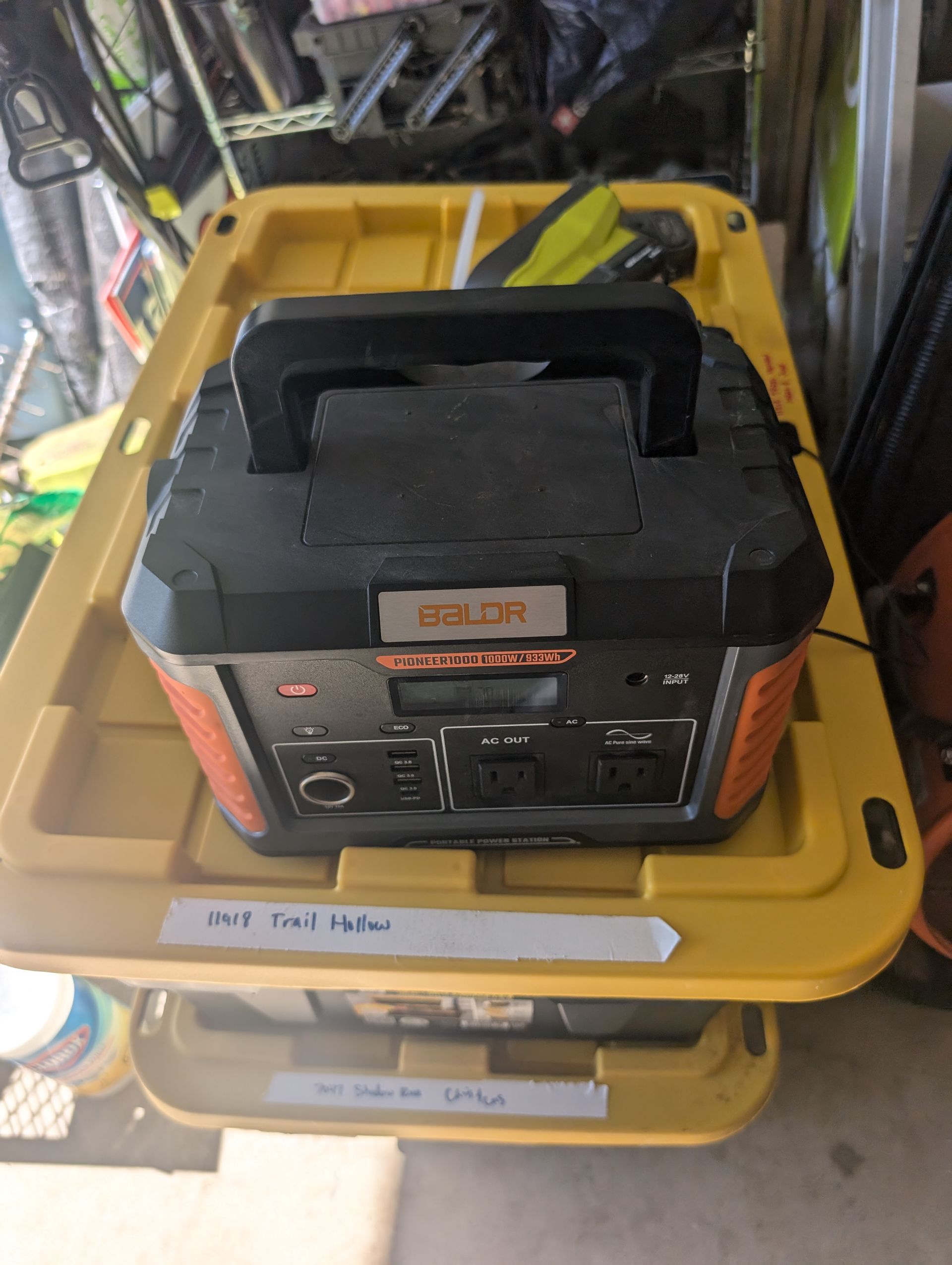 Orange and black portable power station with a black handle, sitting inside a yellow storage bin.