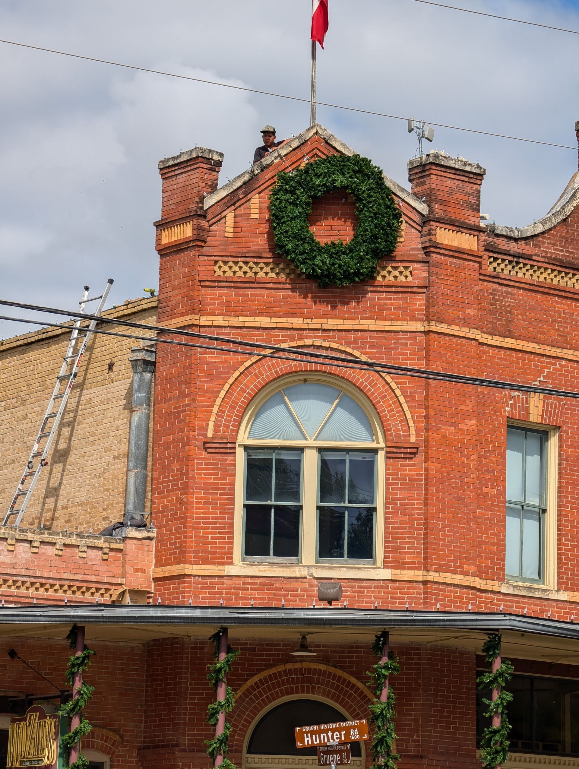 Red brick building with a green wreath and a flag on top. A window with an arched top is below.