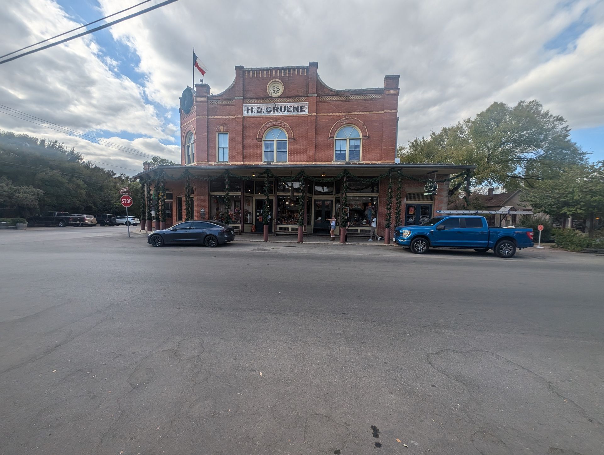 Historic red brick building with a porch, storefronts, and parked vehicles on a street corner.