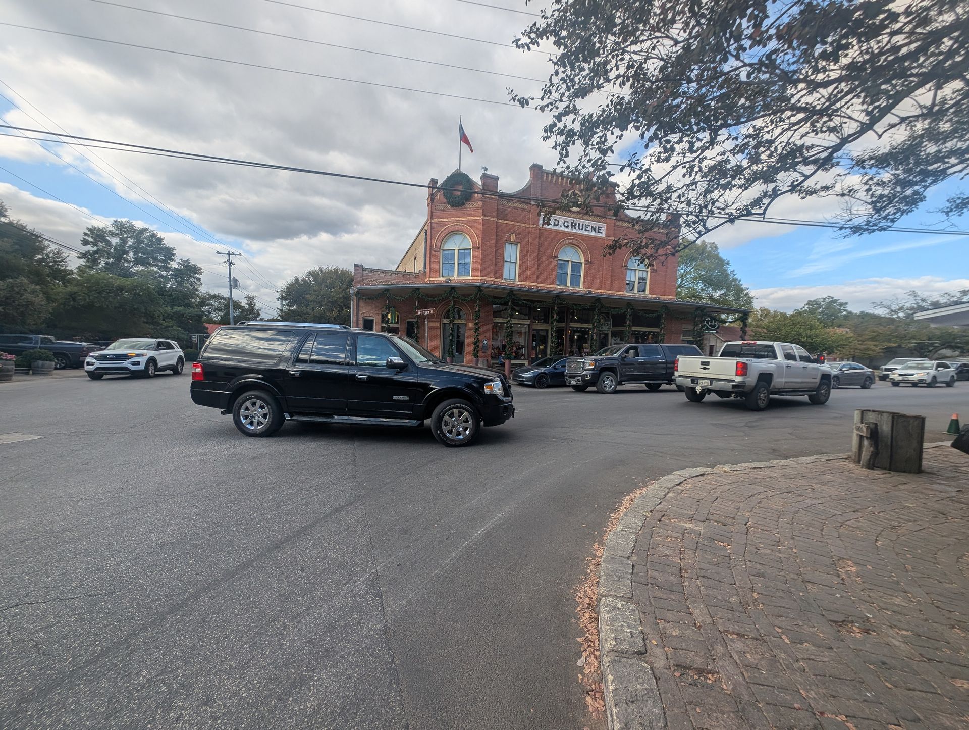 Black SUV driving in front of a brick building with a group of cars parked. Cloudy day.