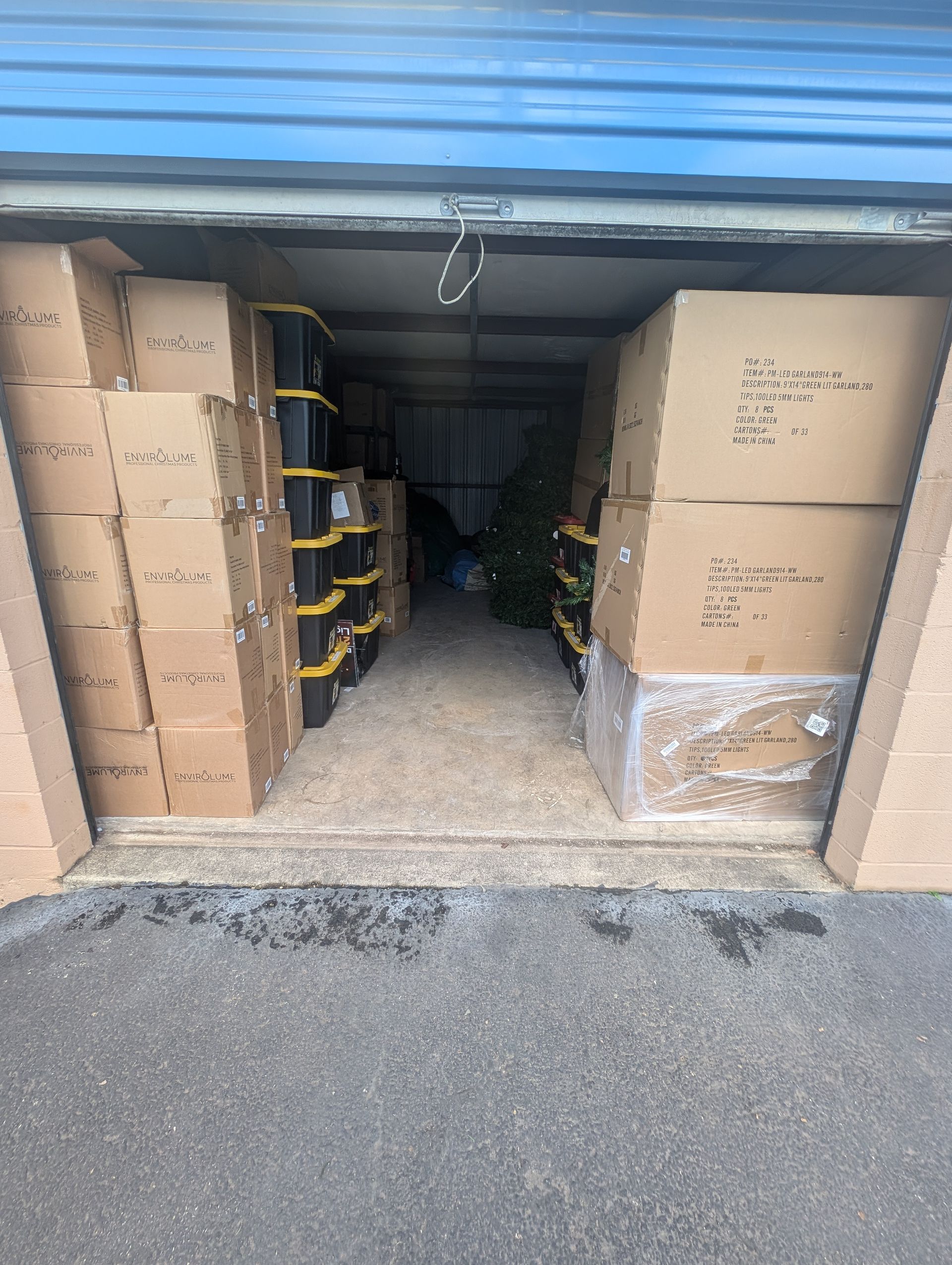 Storage unit filled with boxes, some stacked, and black bins. Door open, showing items inside.