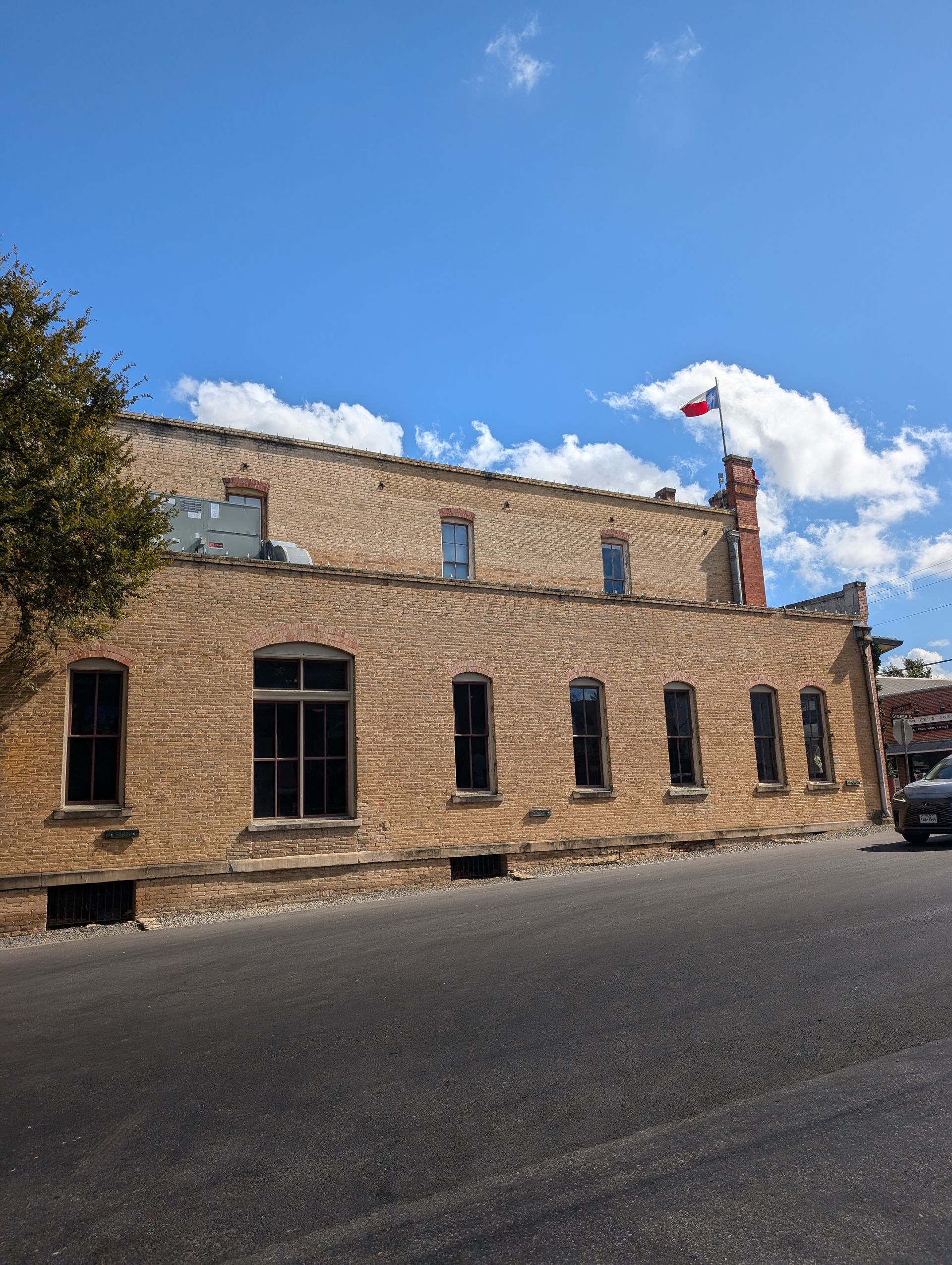 Brick building with Texas flag on a sunny day. Dark windows, a chimney, and a road in the foreground.