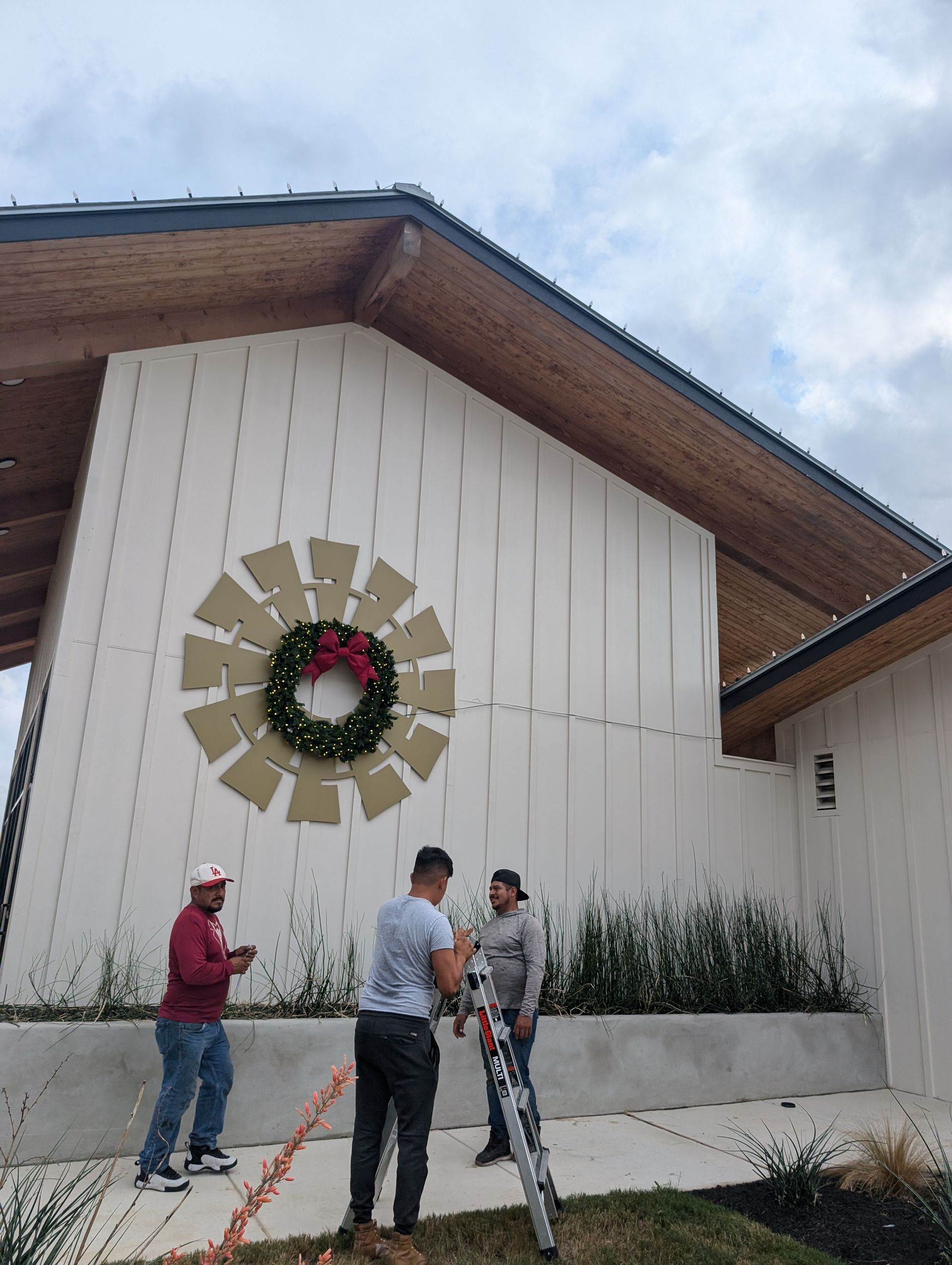 Three men decorate a white building with a wreath; wood trim and a neutral-colored windmill feature.