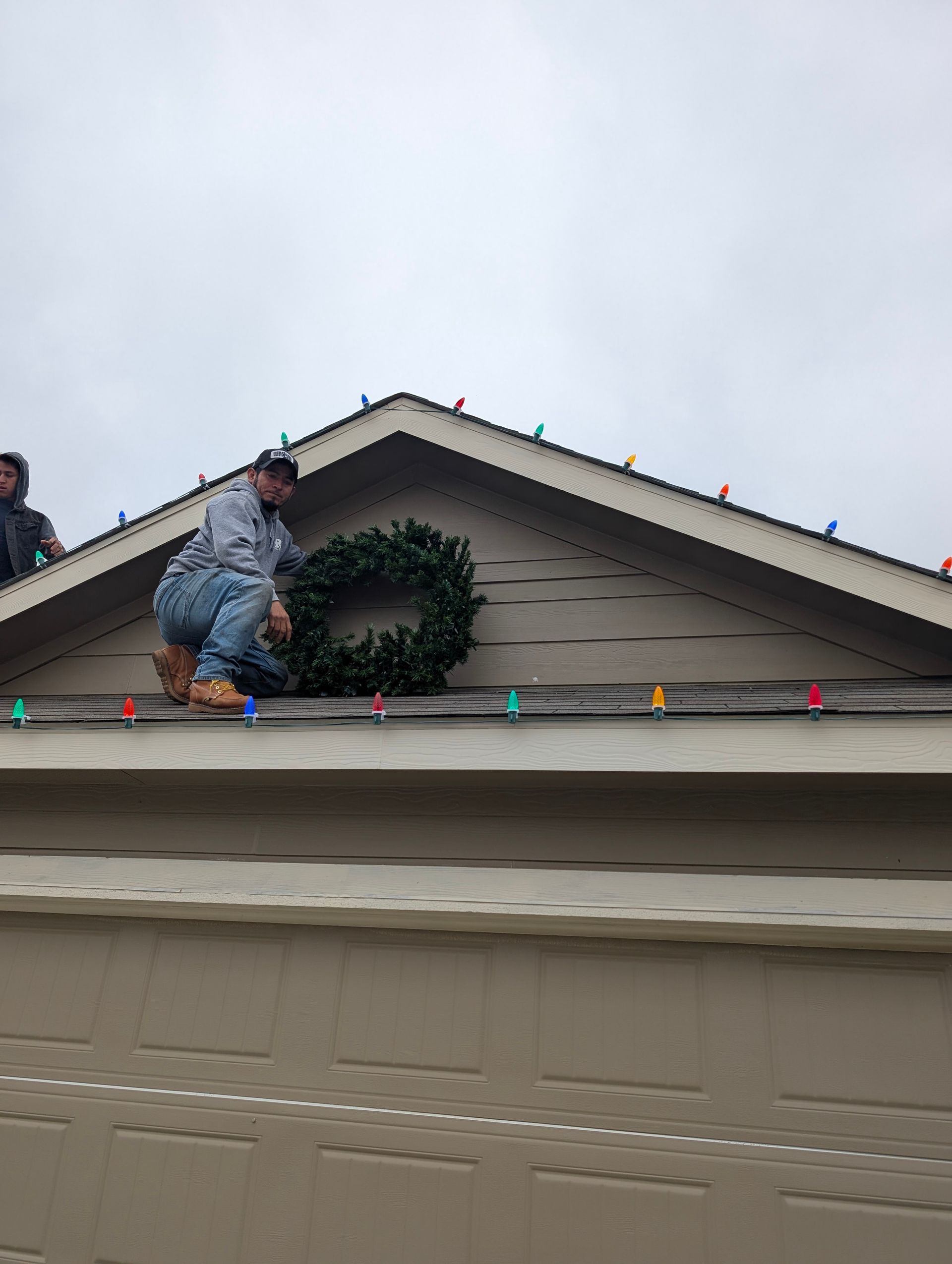 Man on roof, installing a Christmas wreath. Beige house, colorful lights, overcast sky.